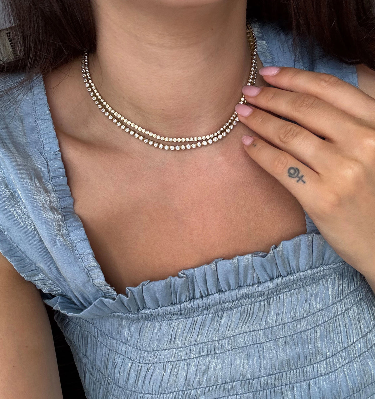 Woman wearing a gold necklace with multiple rows of circular diamonds, dressed in a light blue top.