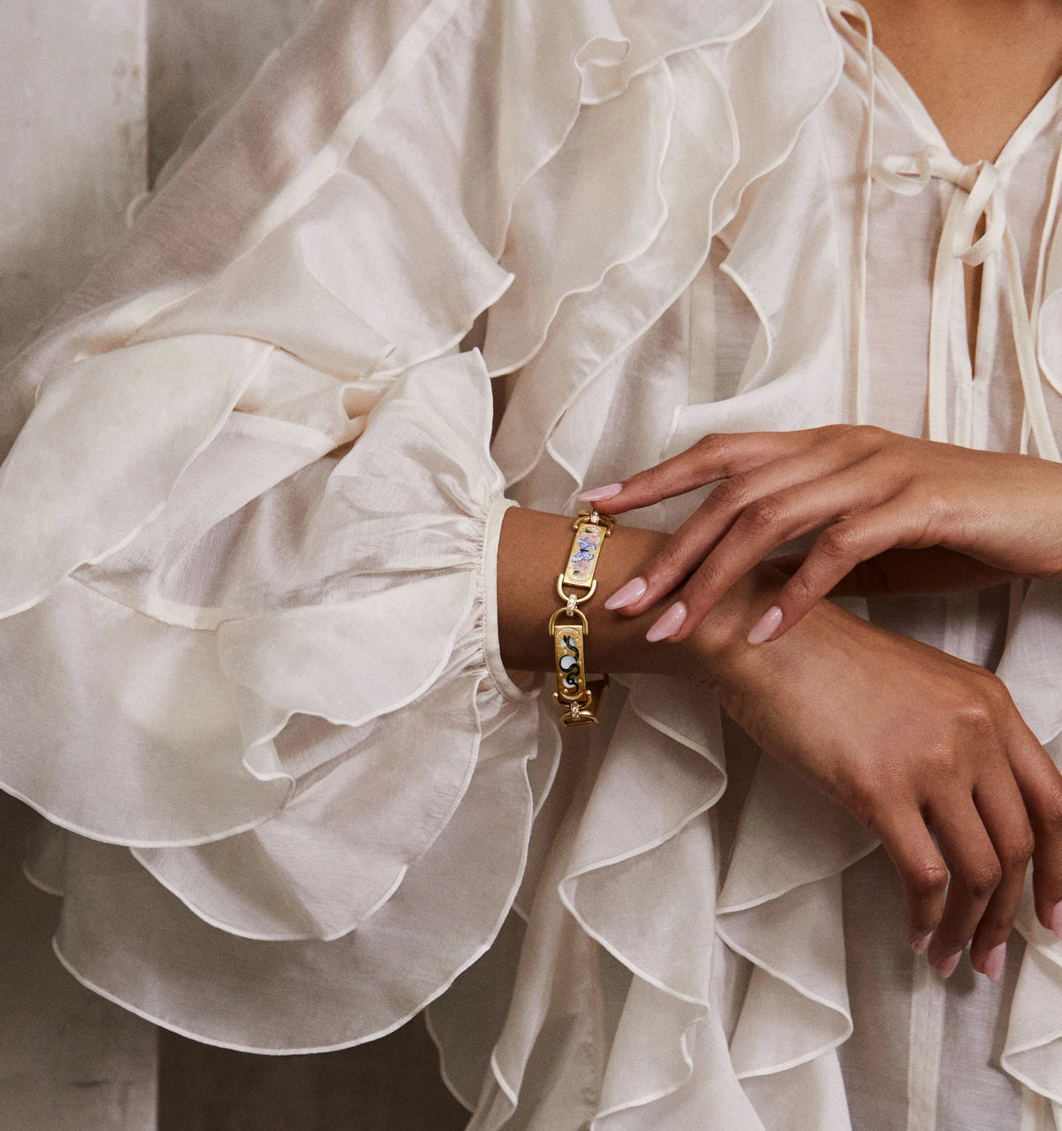 Woman wearing a gold bracelet with colourful enamel accents, paired with a ruffled white blouse.
