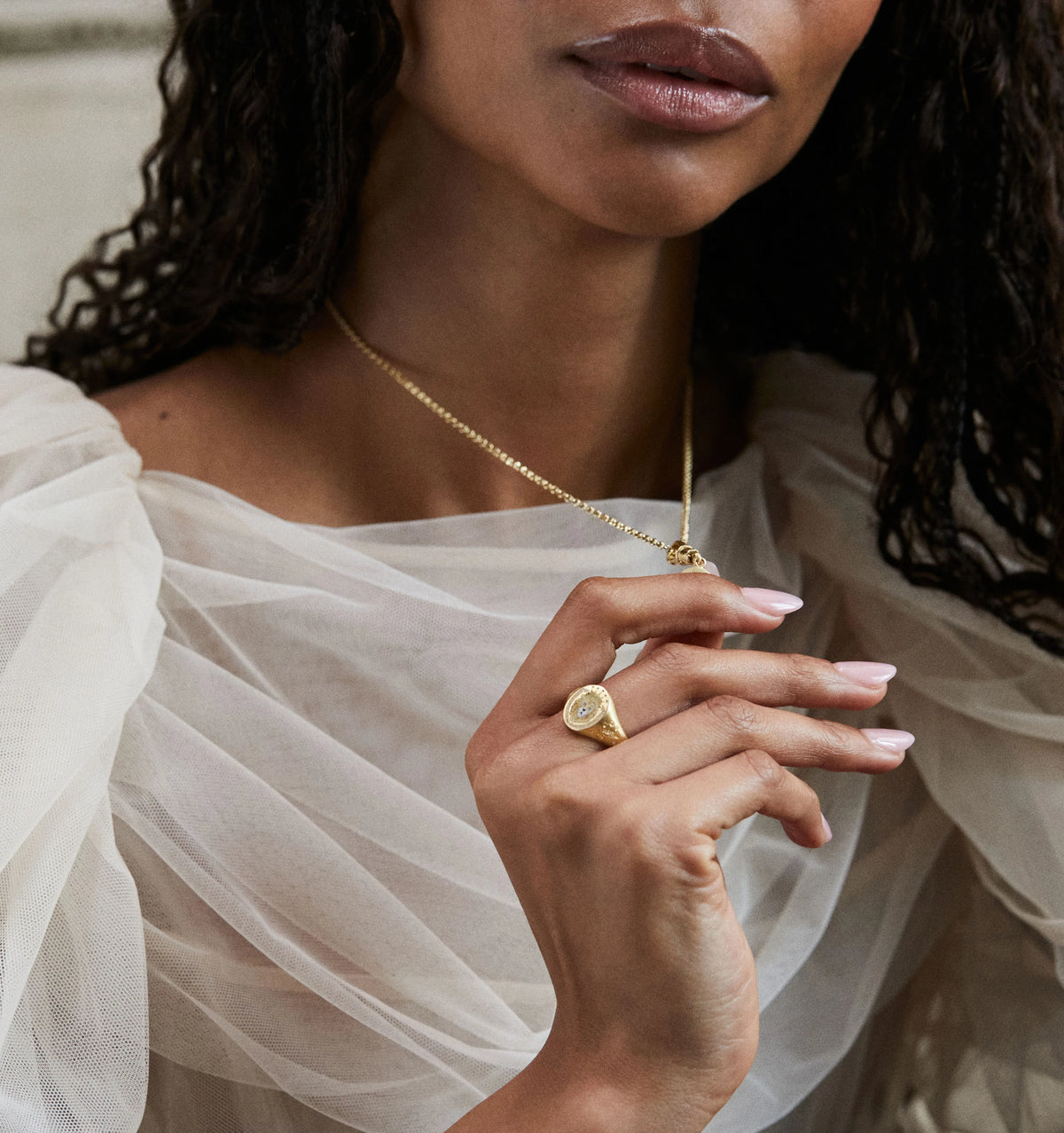 Woman wearing a gold pendant necklace and gold ring with a round design, styled with a flowing sheer top.
