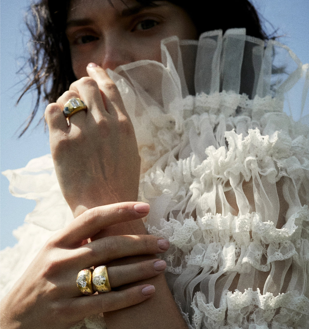 Woman wearing two gold rings with diamonds, posing with hands near her face against a bright background.