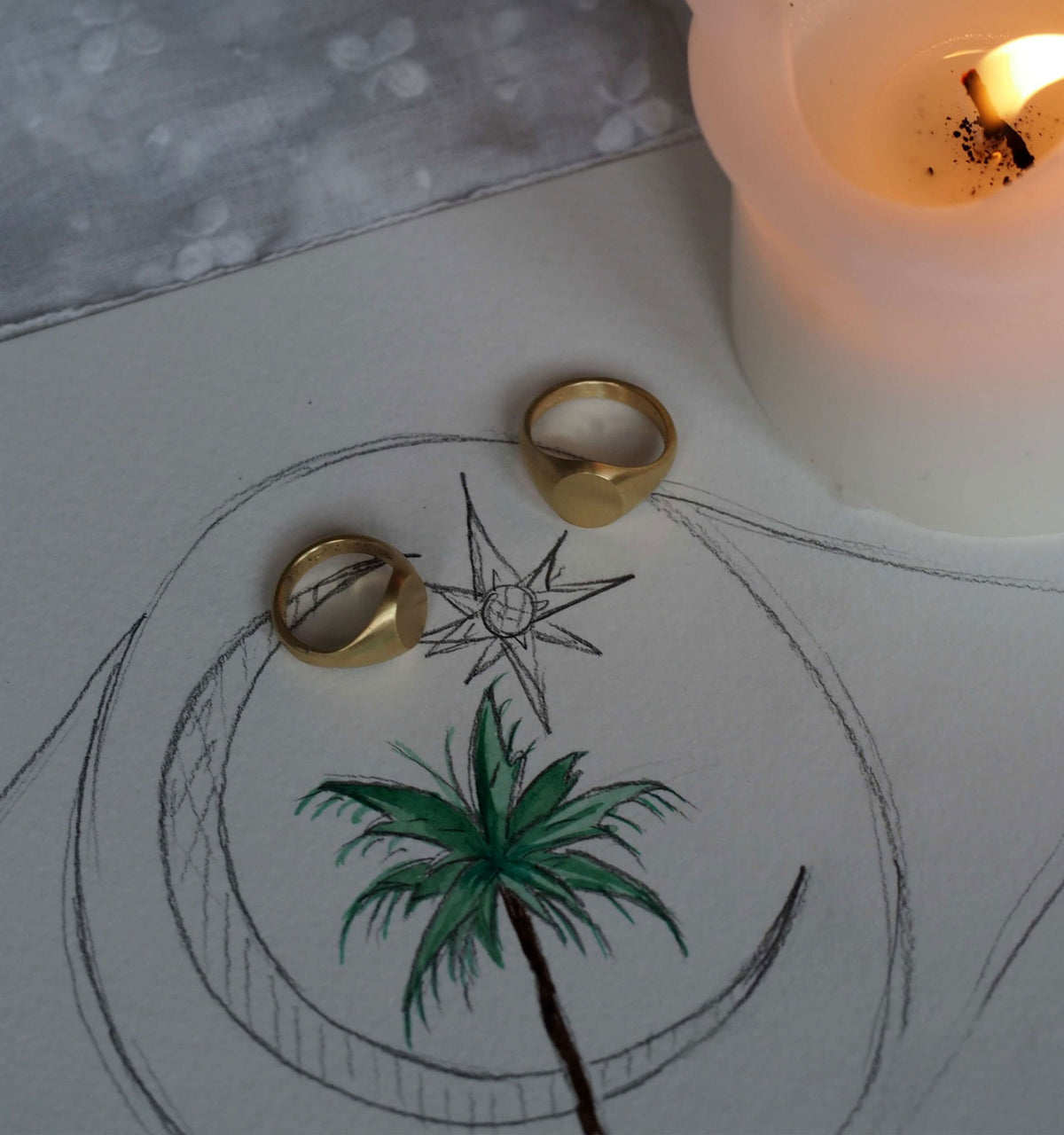 Gold rings placed on a drawing featuring a palm tree and a star, alongside a lit candle.