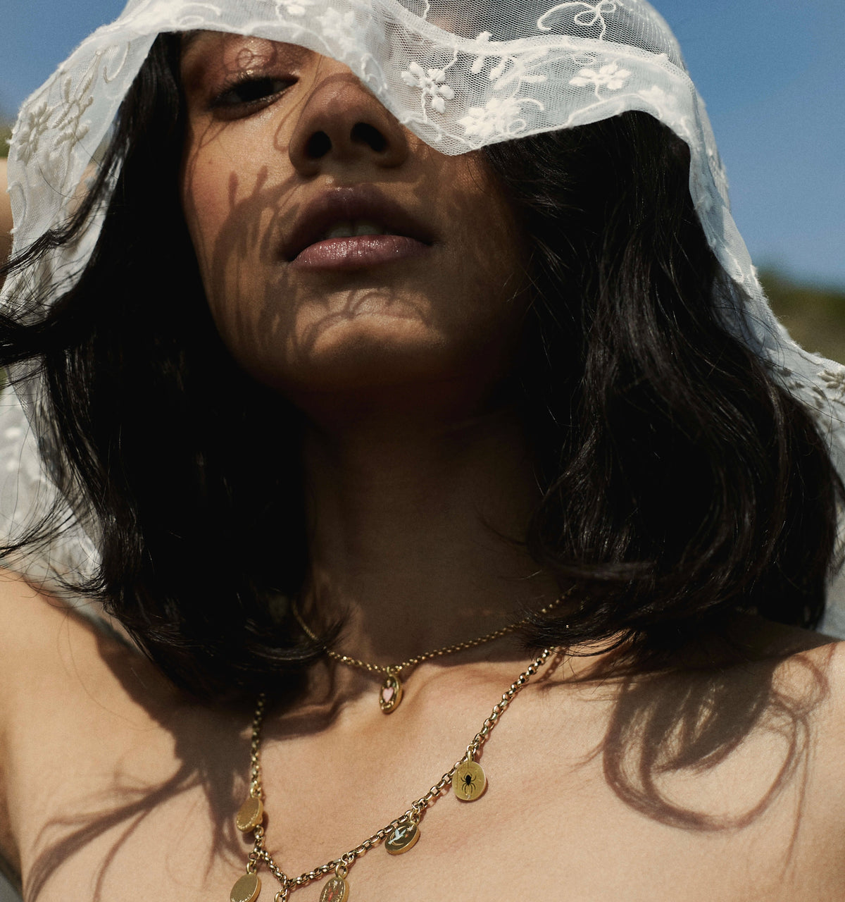 Woman wearing a gold necklace with pendants, partially covered by a sheer veil against a bright outdoor backdrop.