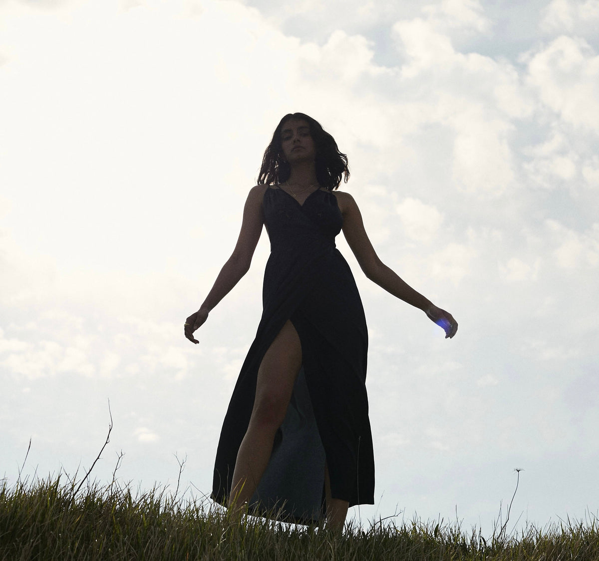 Woman wearing gold jewellery, standing in a field against a cloudy sky, dressed in a flowing black gown.
