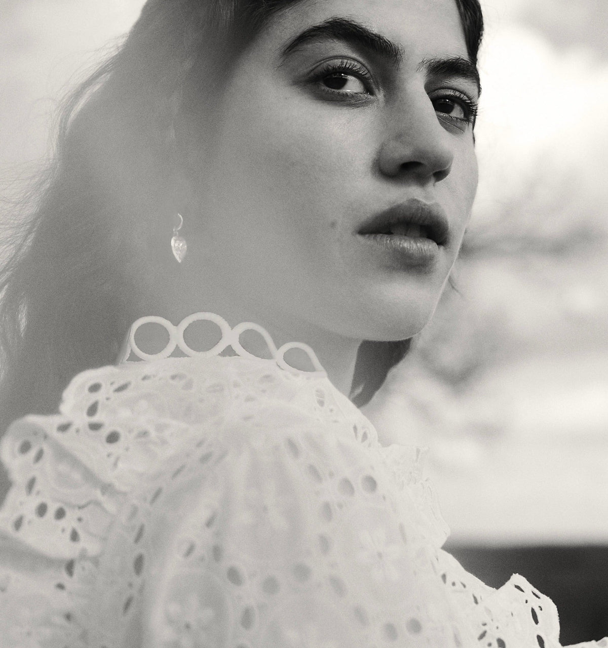 Woman wearing gold earrings and a detailed white blouse, set against a soft-focus background.