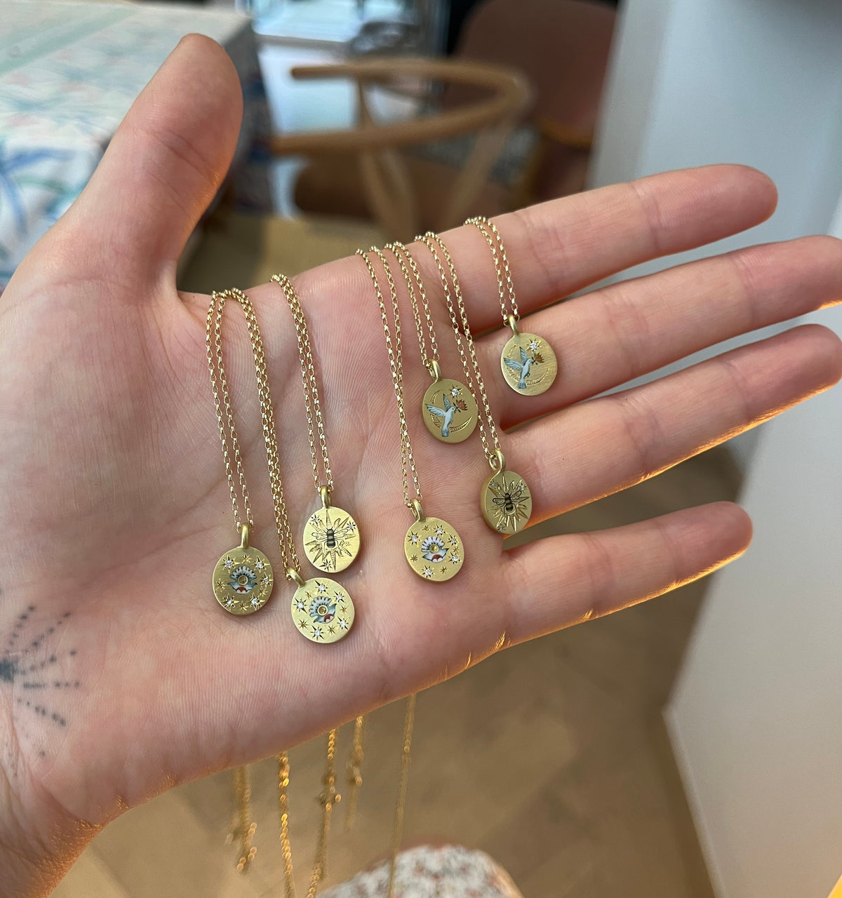 Gold necklaces featuring engraved pendants with colourful motifs, held in a hand against a soft background.