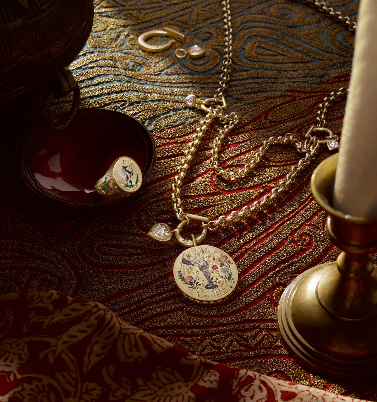 Gold pendant necklace with floral enamel design, gold ring, and gold hoop earrings on textured red and gold fabric.
