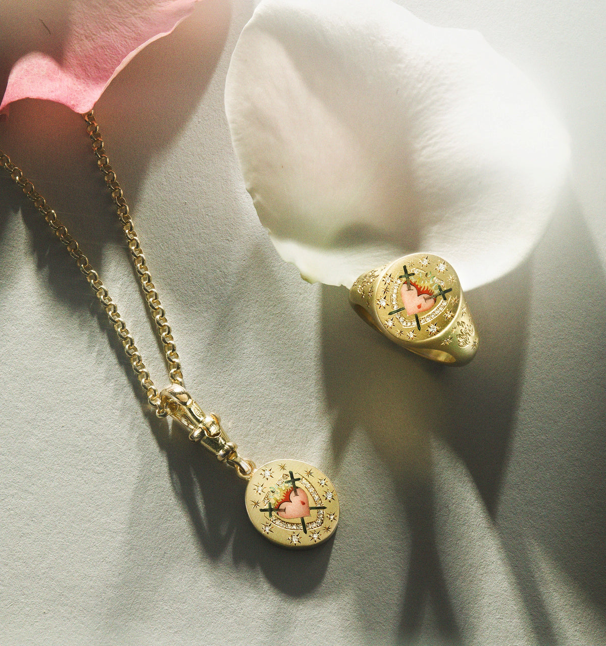 Gold pendant with an engraved heart design and a matching gold ring featuring a heart motif, displayed on soft petals.