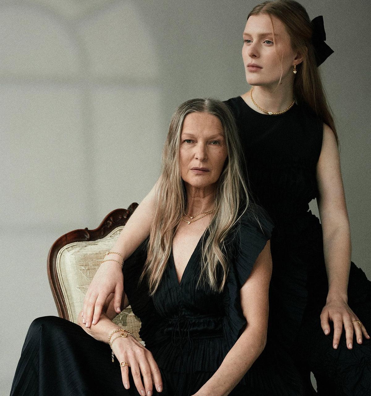 Two women wearing gold jewellery, including layered necklaces, bracelets, rings, and drop earrings, in black dresses.