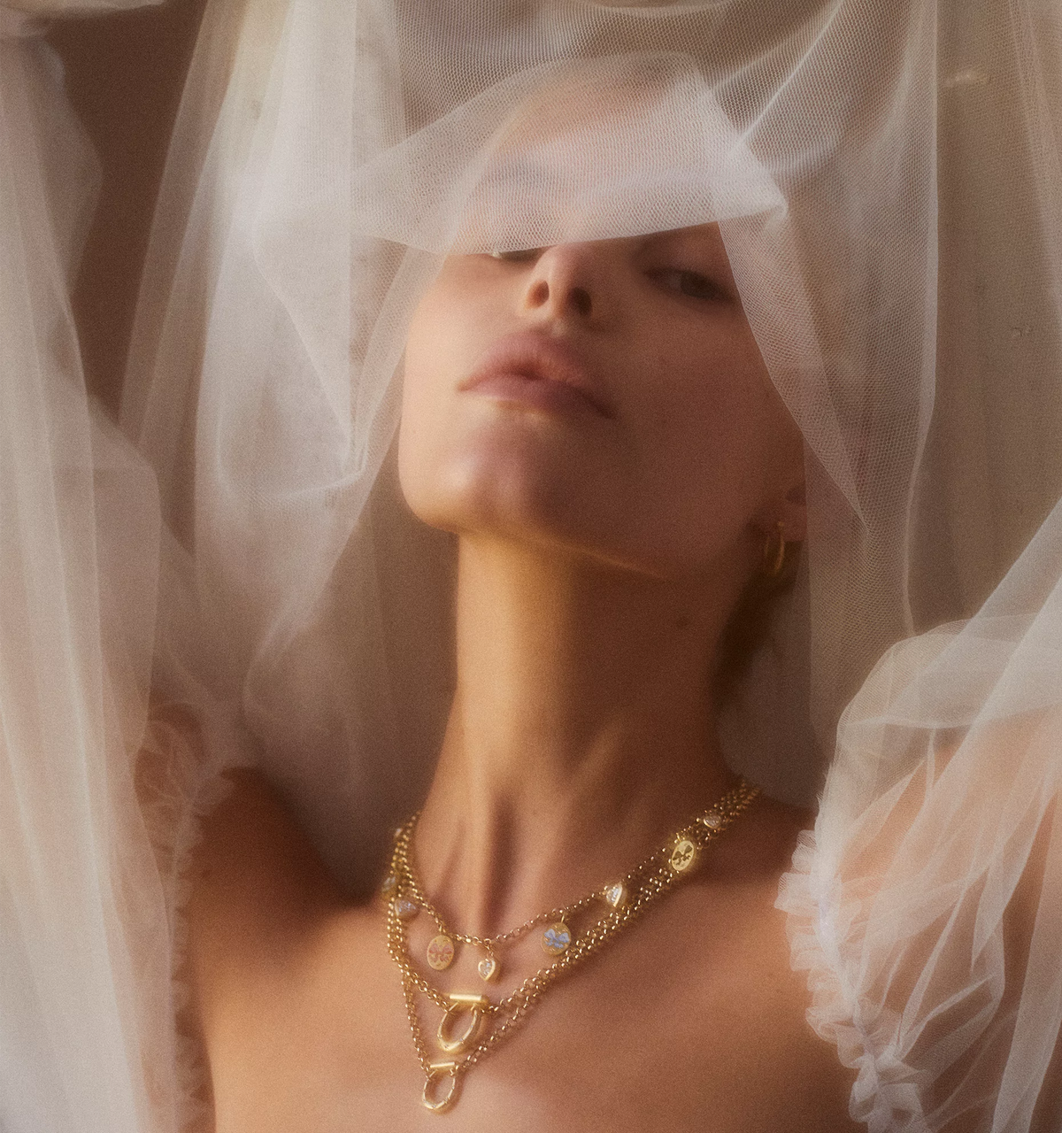 Woman wearing layered gold necklaces with heart and oval charms, veil draped softly over her face.