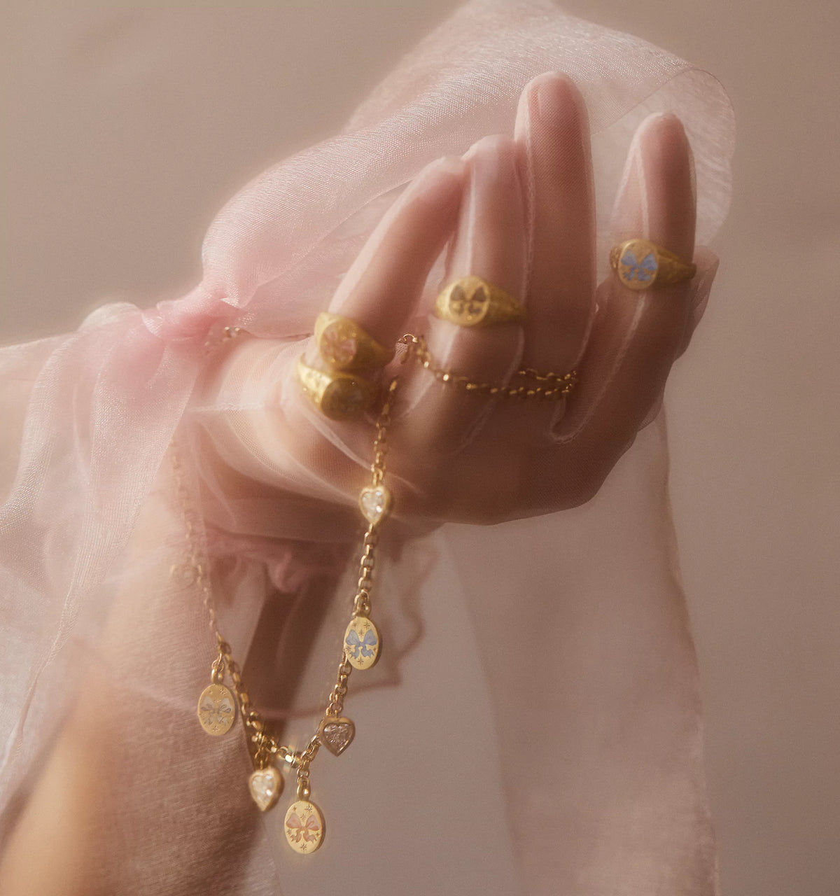 Woman wearing gold signet rings with enamel bow motifs, holding a necklace with heart and bow charms, through sheer fabric.