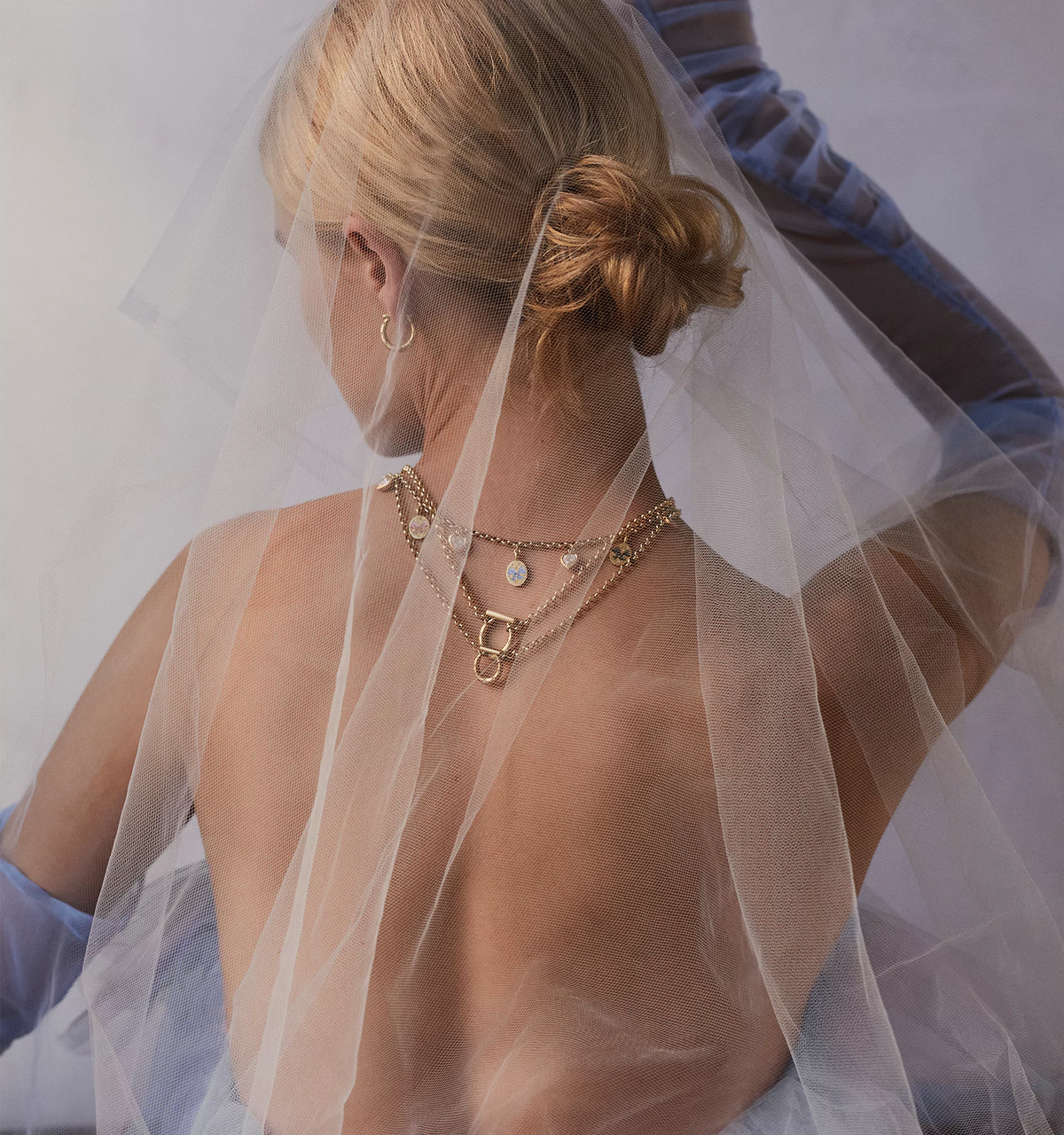 Woman wearing layered gold necklaces with pendant and details, partially hidden by a sheer veil.