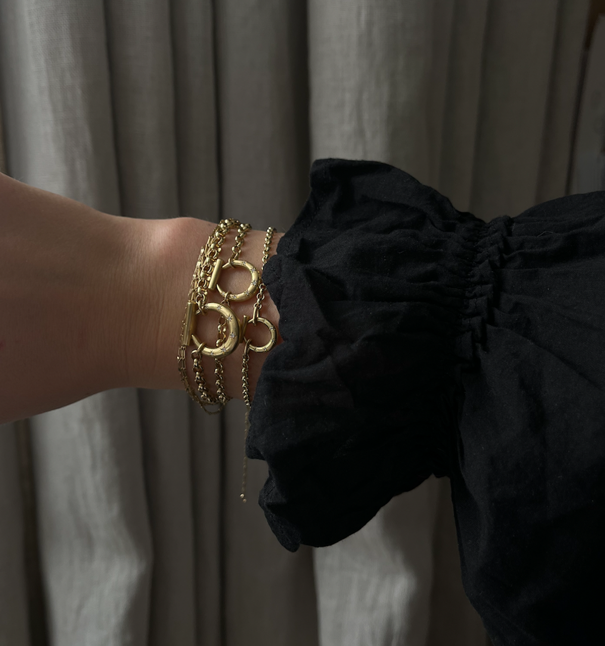 Woman wearing multiple gold bracelets against a backdrop of fabric drapes.
