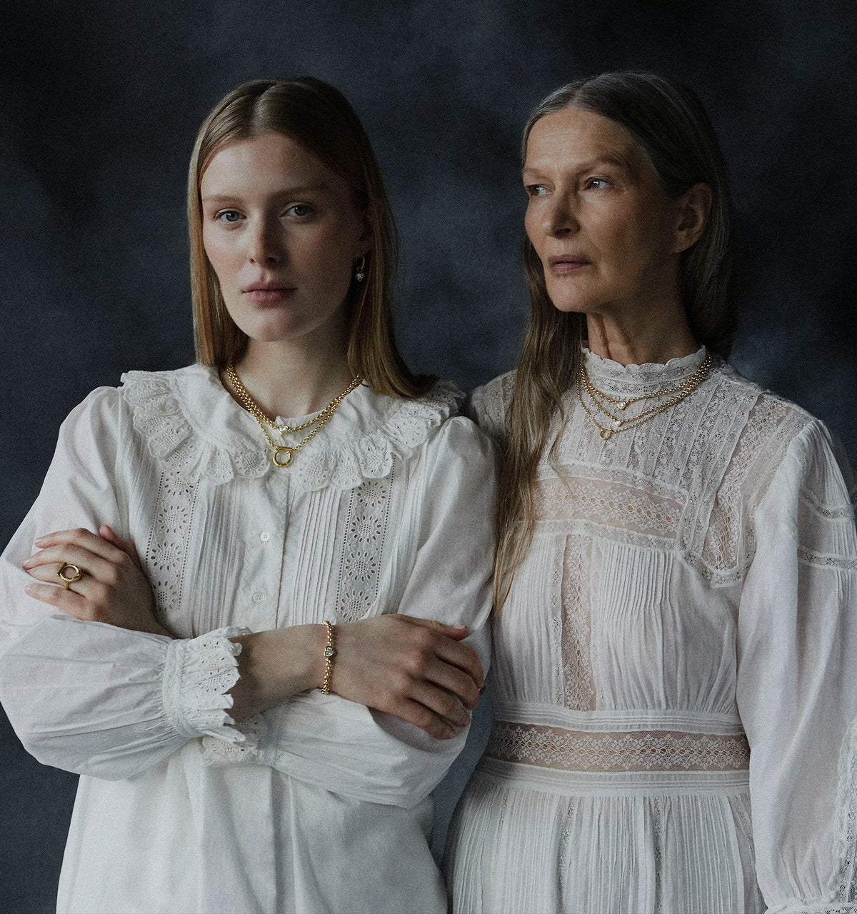 Woman wearing layered gold necklaces and gold earrings, alongside another woman in a white lace-trimmed blouse.