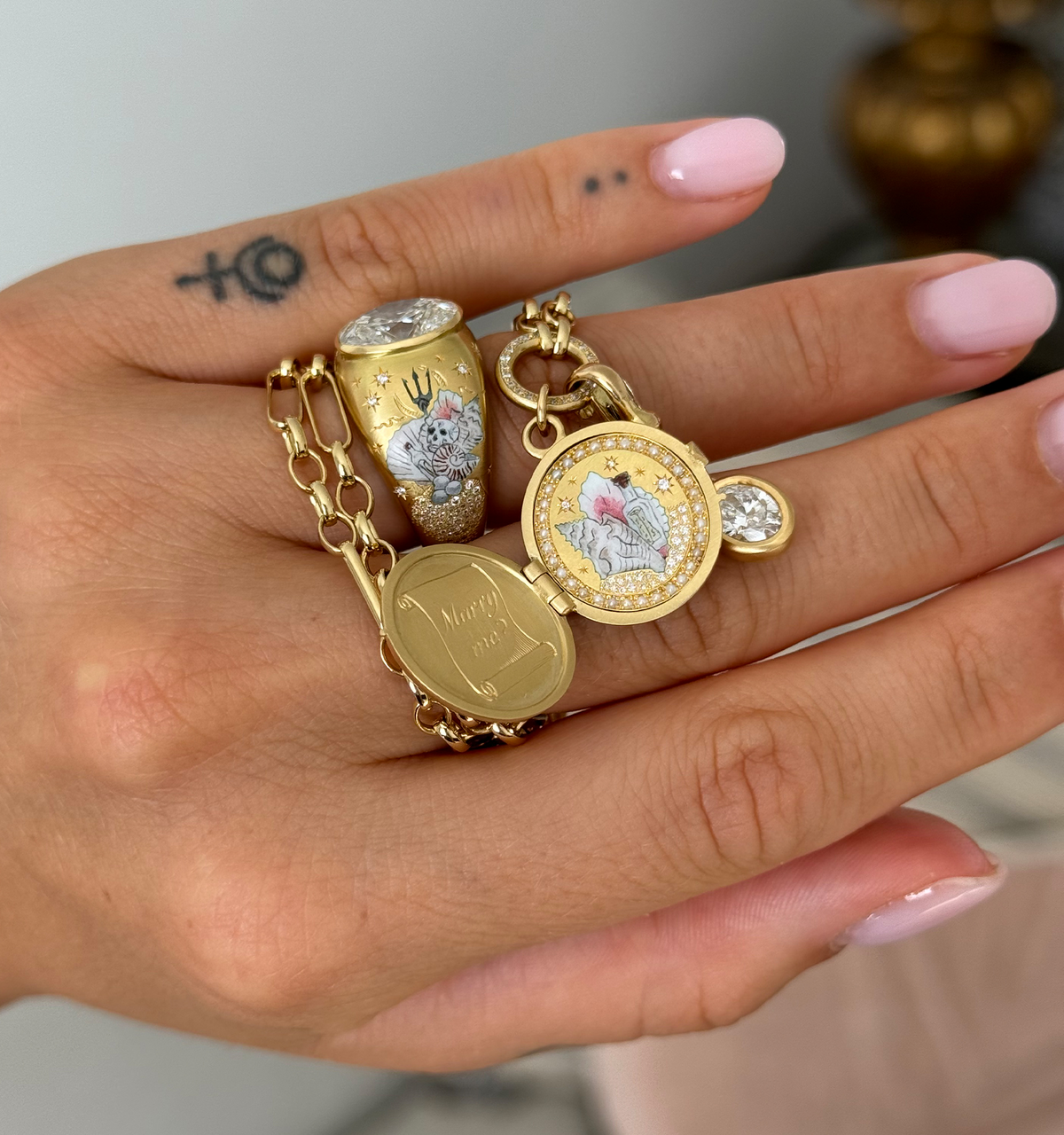 Gold bracelet and locket featuring enamel designs with intricate detailing, worn on a hand with a tattoo.