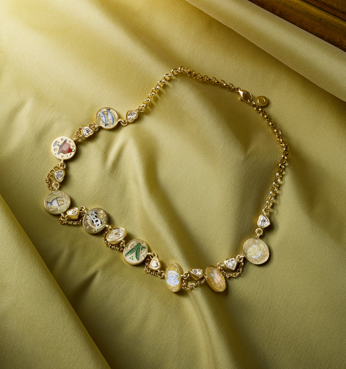 Gold necklace adorned with colourful enamel charms on a soft yellow silk background.