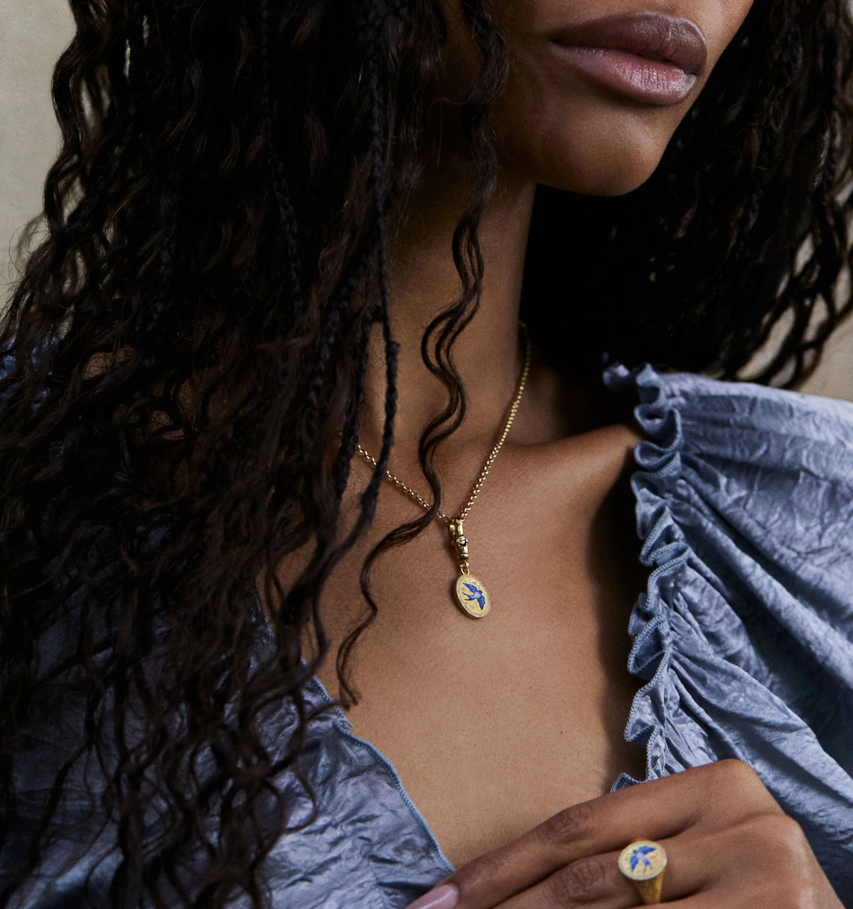 Woman wearing a gold pendant necklace with a blue floral design and matching gold ring.