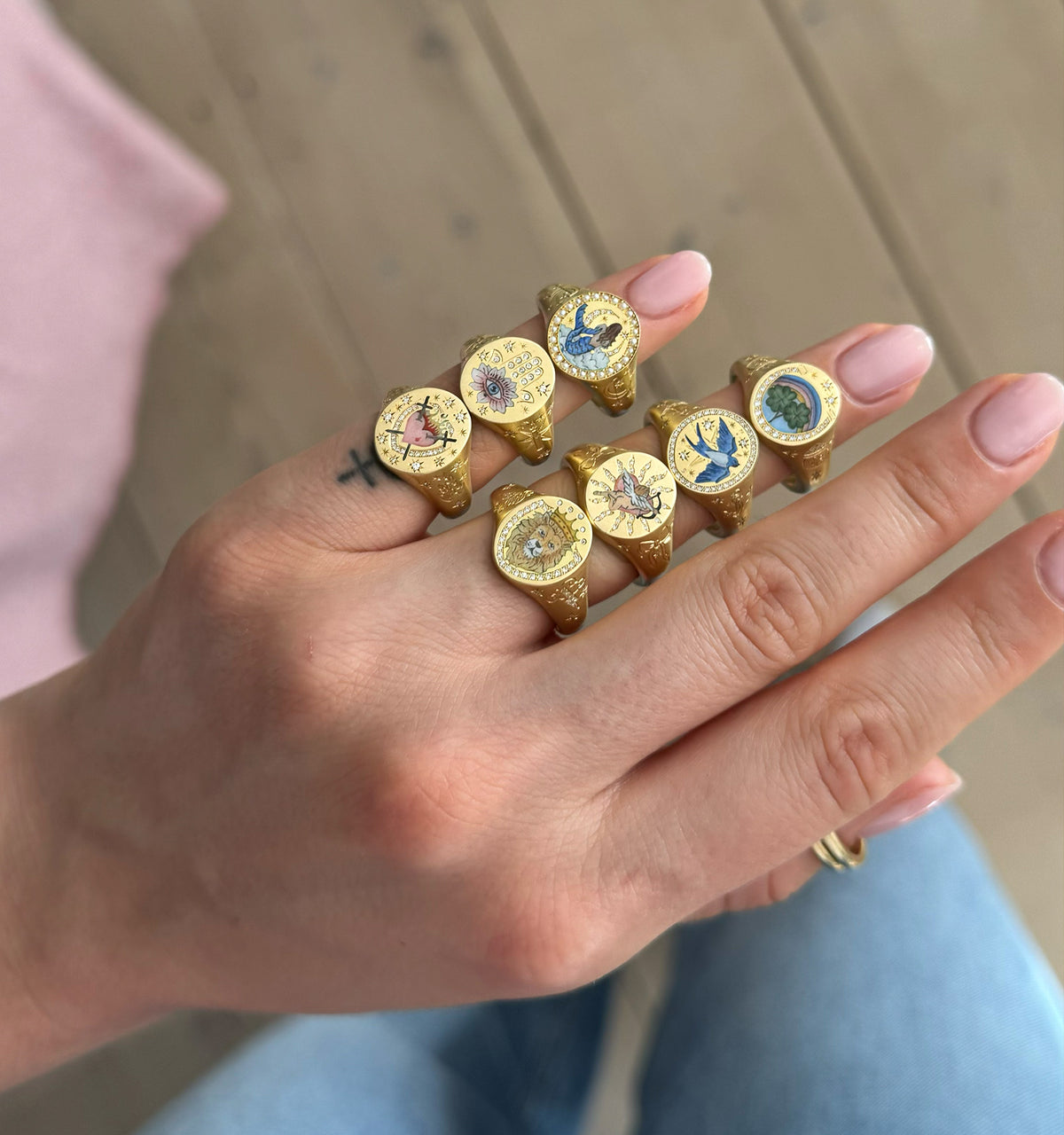 Woman wearing six gold rings with colourful enamel designs, displayed on her hand against a light wooden background.