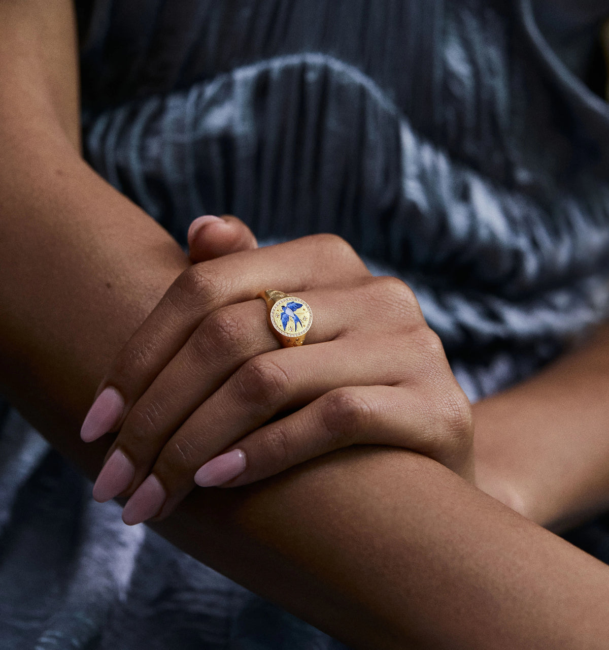 Woman wearing a gold ring featuring a blue design on a textured background, hands crossed with pink nails.