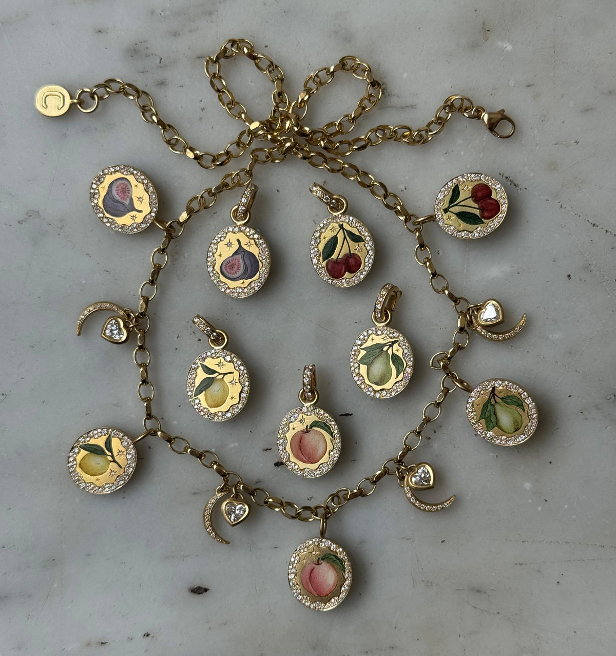 Gold charm necklace featuring enamel fruit motifs and diamonds, arranged on a light-coloured surface.