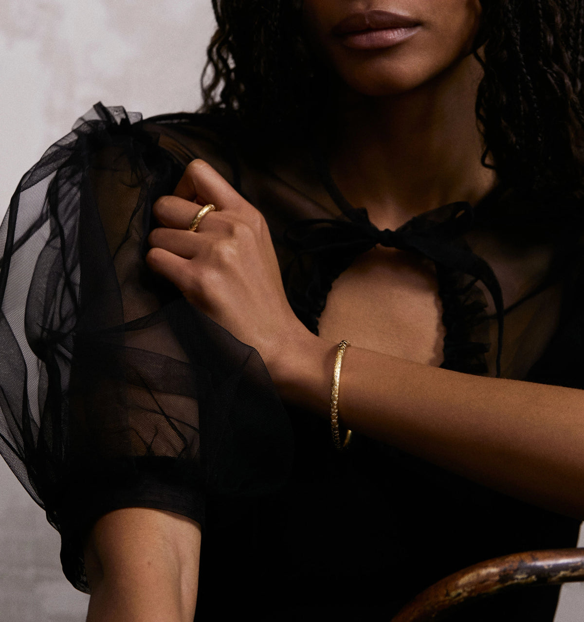 Woman wearing a gold twisted bangle, with sheer black puff-sleeved top in a soft, neutral setting.