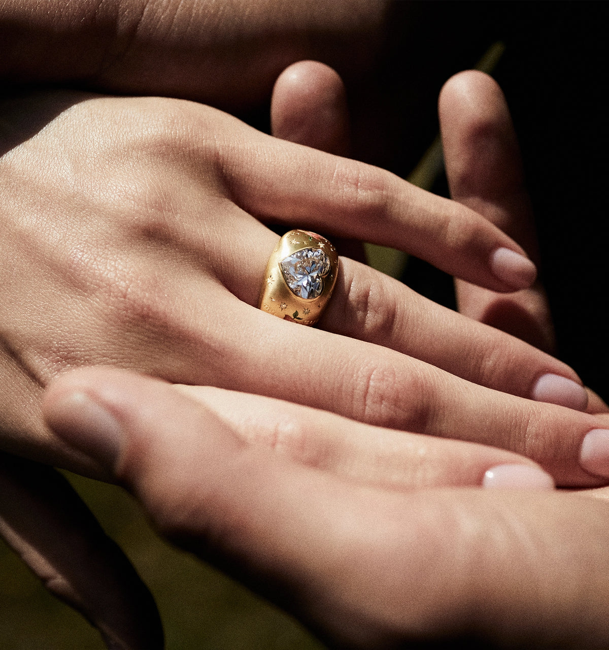 Close-up of a gold ring with a heart-shaped diamond, worn on a hand resting over another hand.