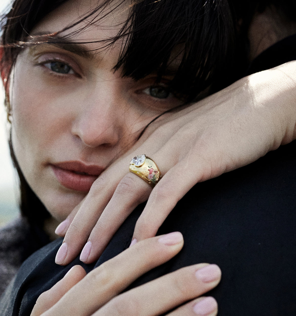 Woman wearing a gold ring featuring a floral motif and a large white stone, resting her hand on another person's shoulder.