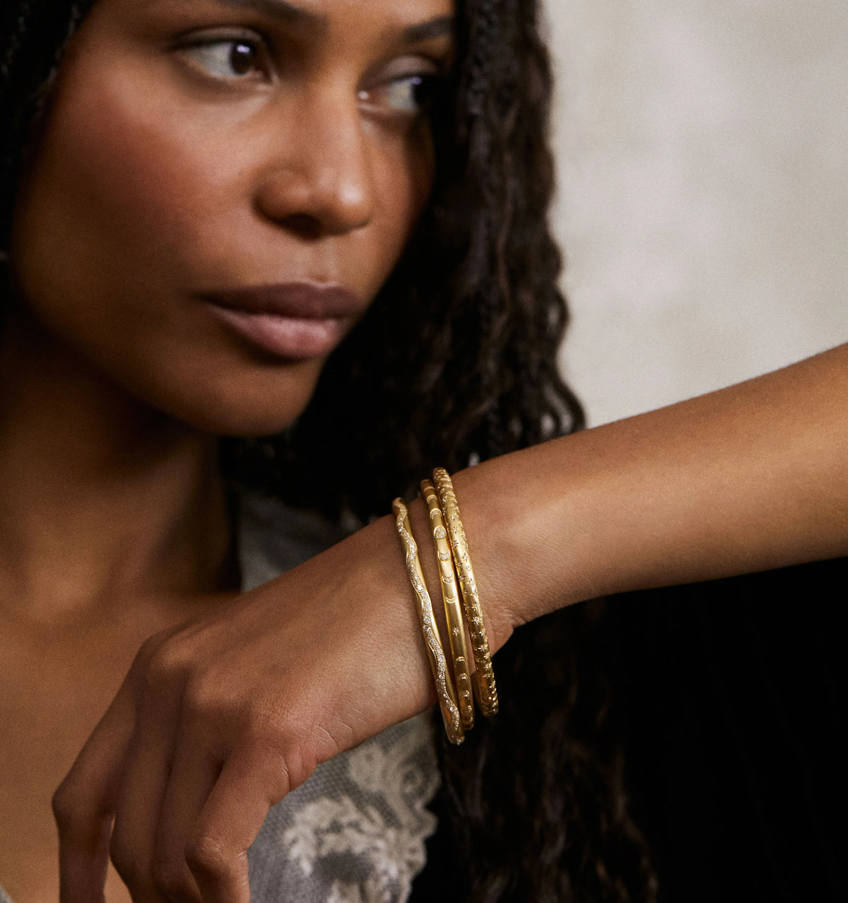 Woman wearing multiple gold bracelets on her wrist against a soft, neutral background.