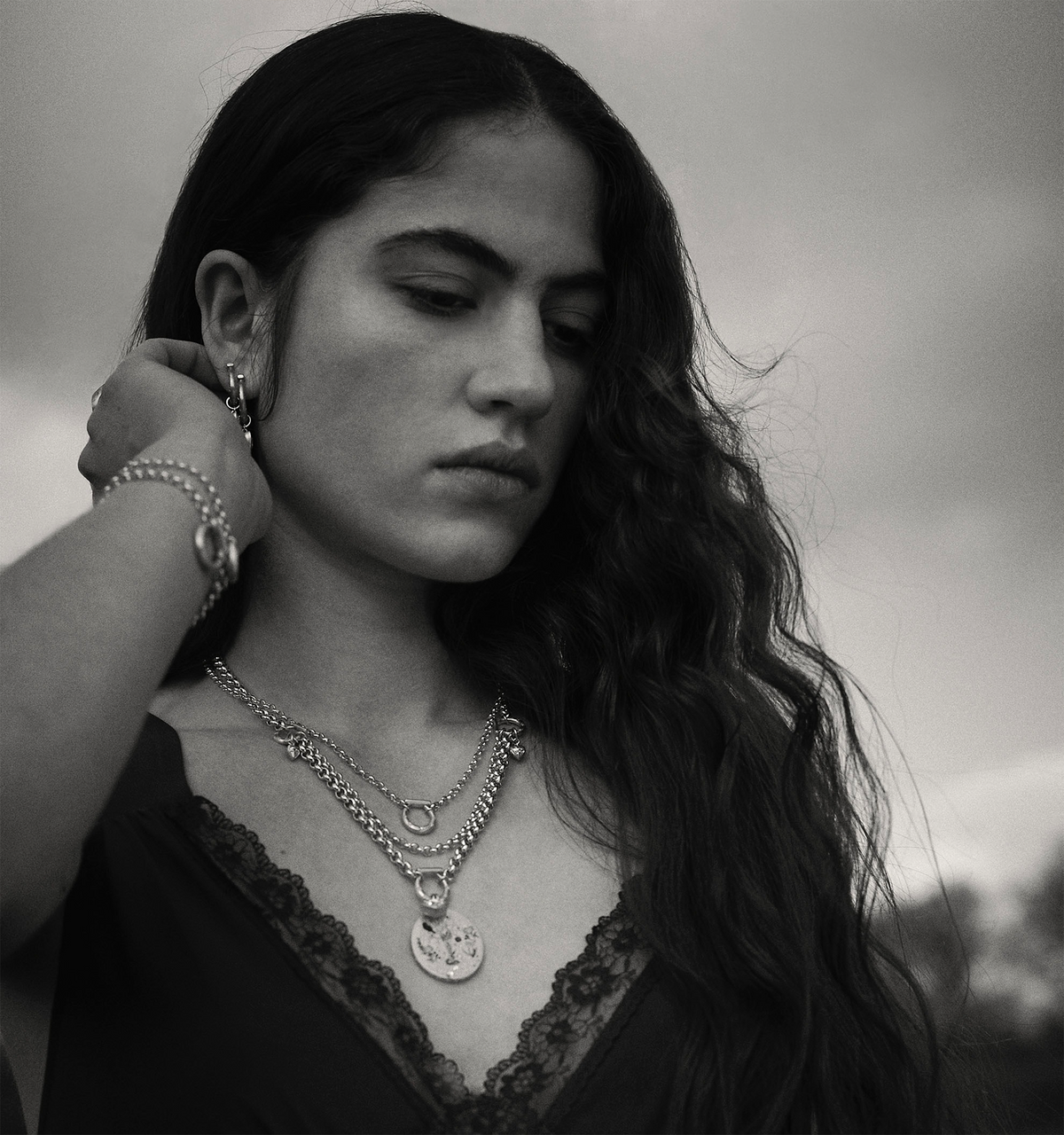 Woman wearing layered gold chains and a gold pendant, with long wavy hair and a black lace top.