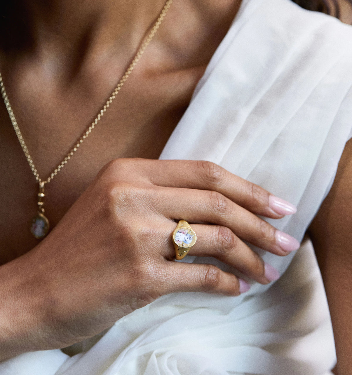 Woman wearing a gold ring featuring a floral design and a gold necklace with a pendant against a light fabric background.