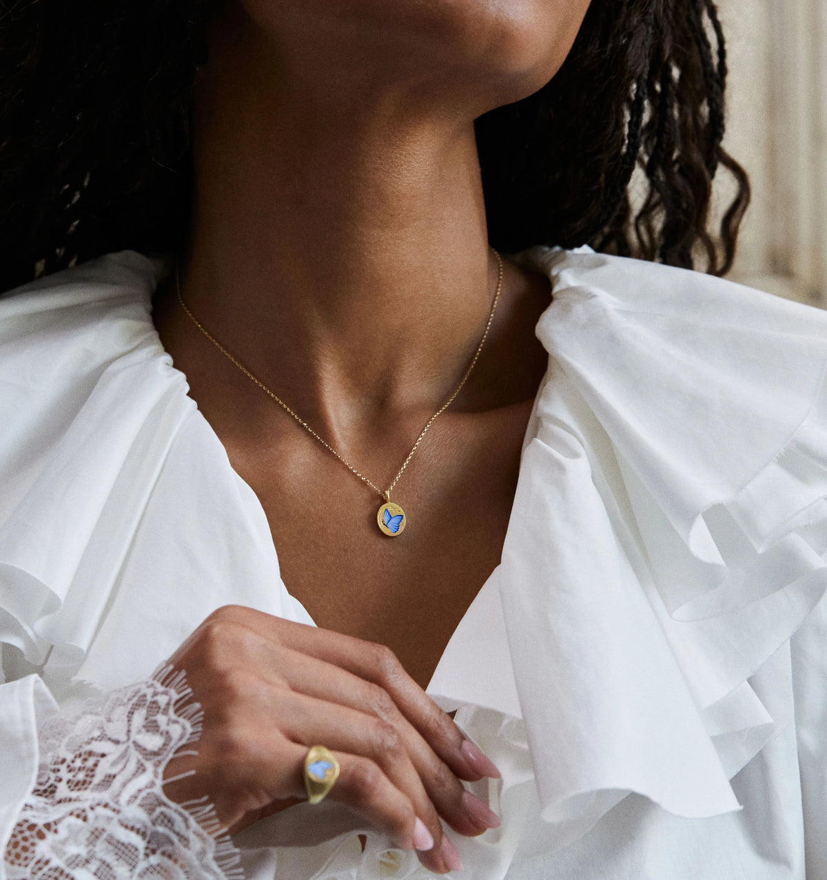 Woman wearing a gold necklace with a blue butterfly motif and a gold ring with an enamel design.