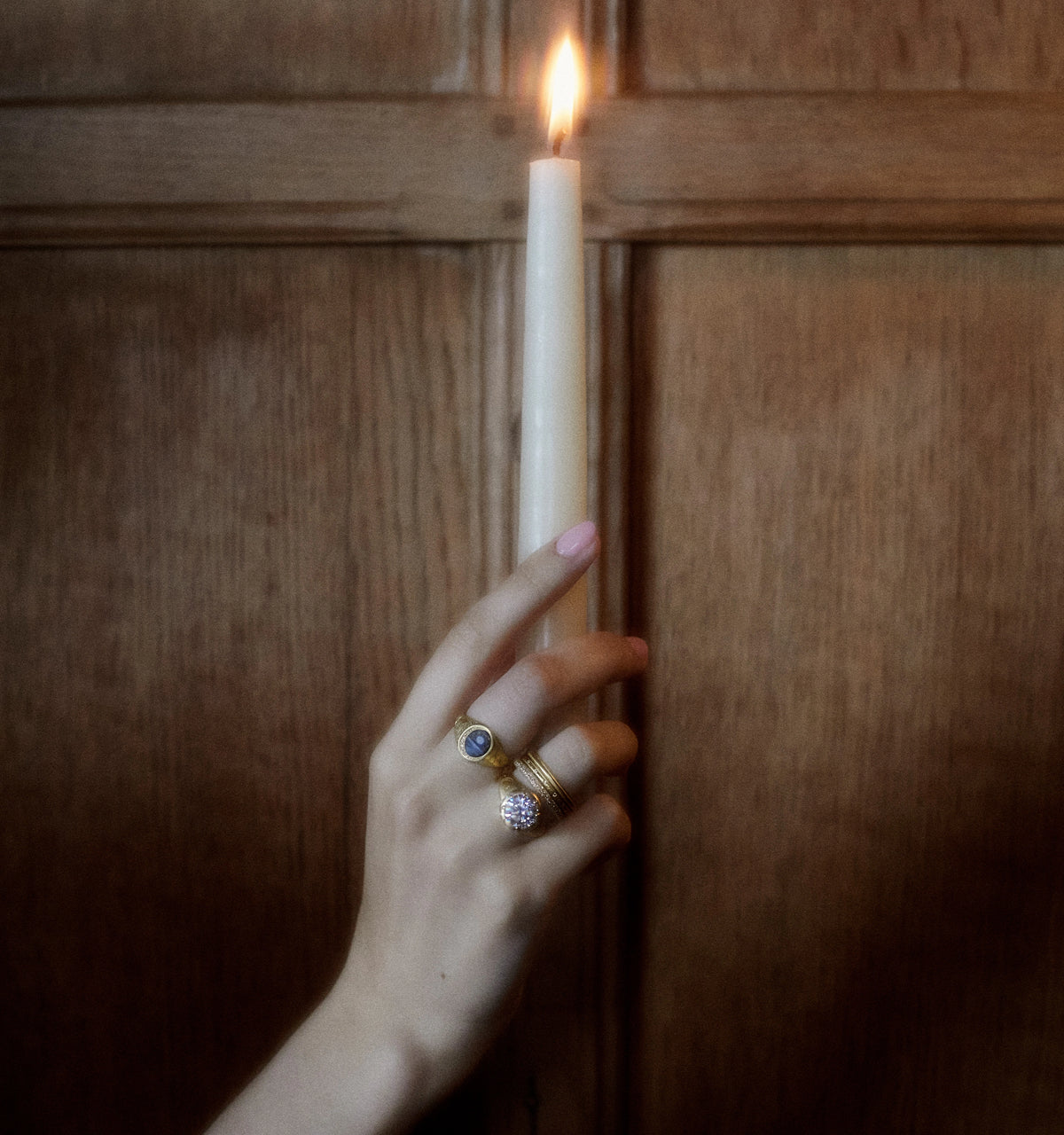 Woman wearing gold rings with a blue stone and a diamond detail, holding a lit candle against a wooden background.