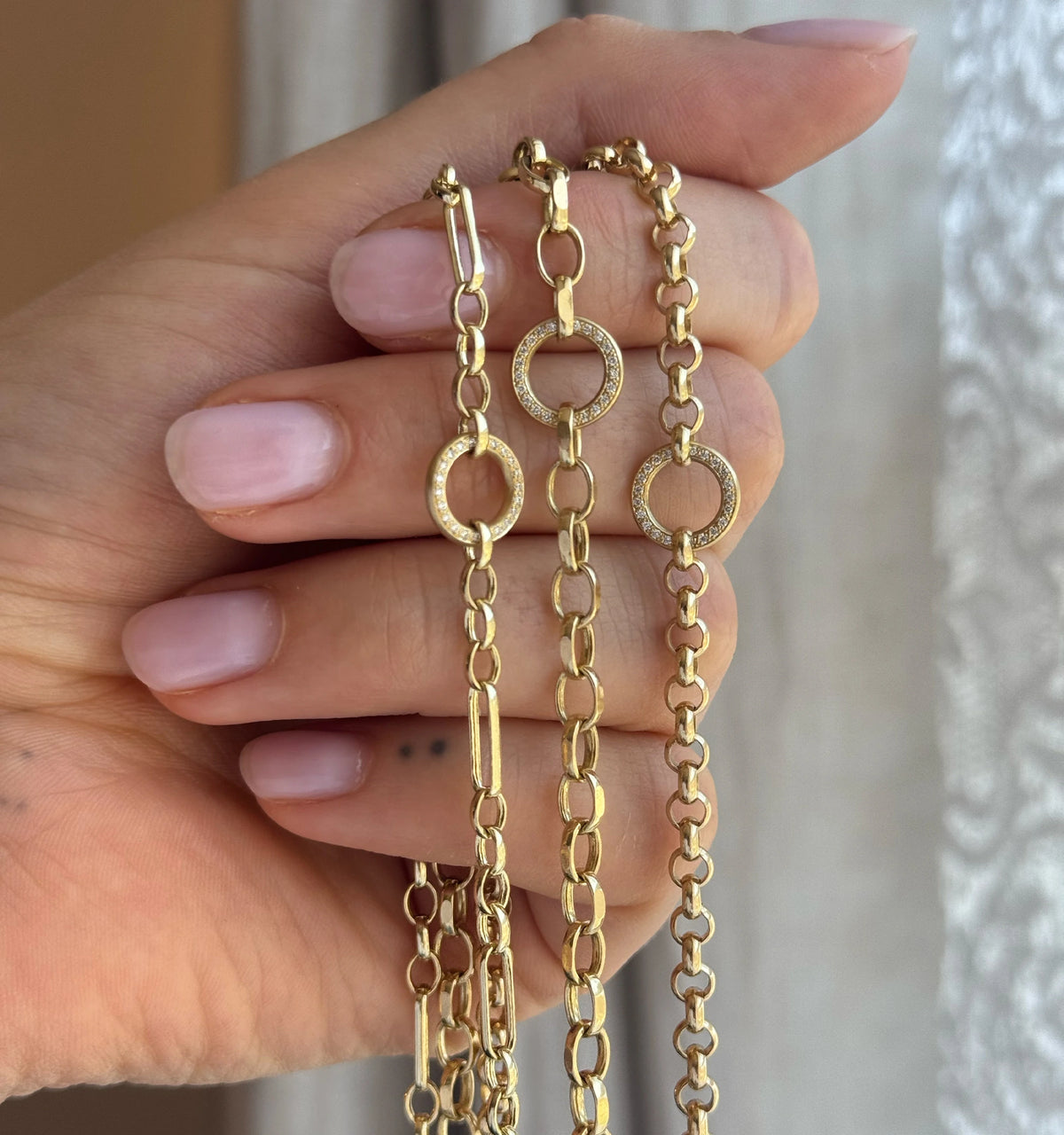 Hand holding three gold chain necklaces, featuring circular links and one with a diamond-encrusted circle.