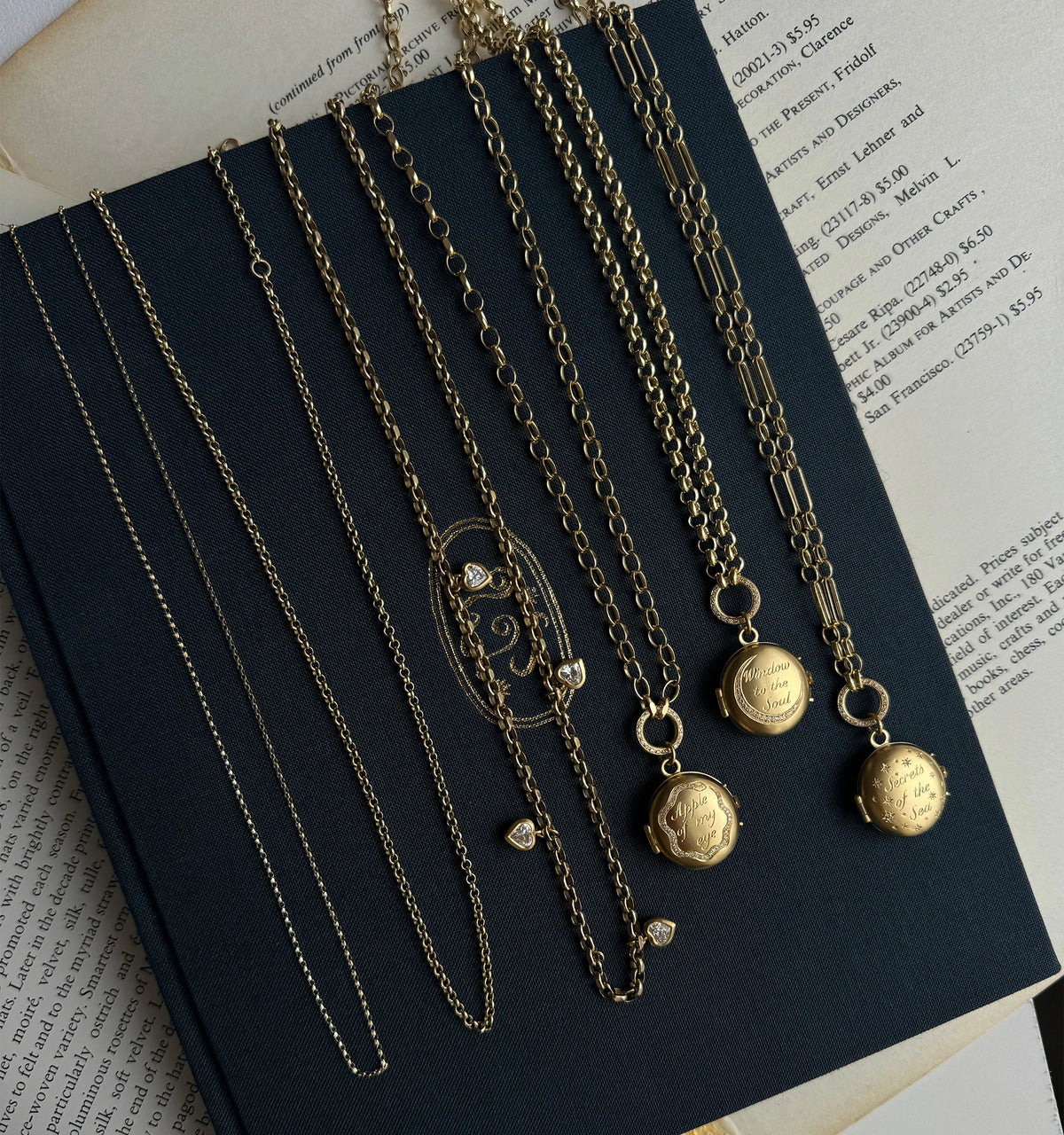 Gold necklaces displayed on an open book, featuring varied chains and two lockets with engraved designs.