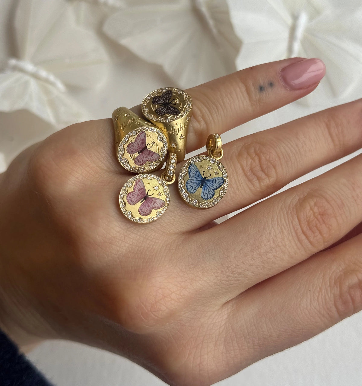 Woman wearing multiple gold rings and a pendant featuring colourful butterfly designs with enamel detailing.