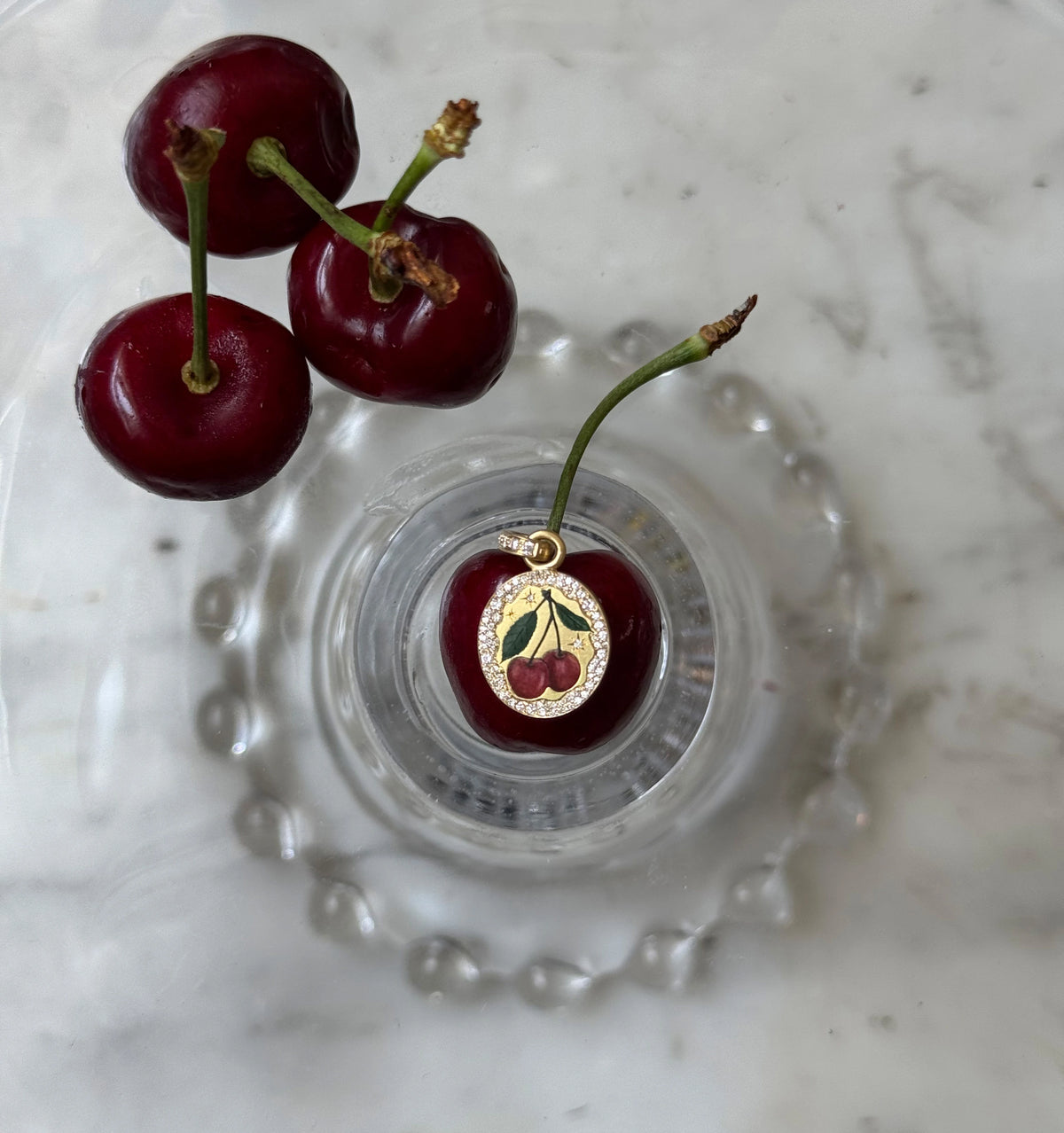 Gold pendant featuring cherries in enamel placed on a glass dish with fresh cherries.