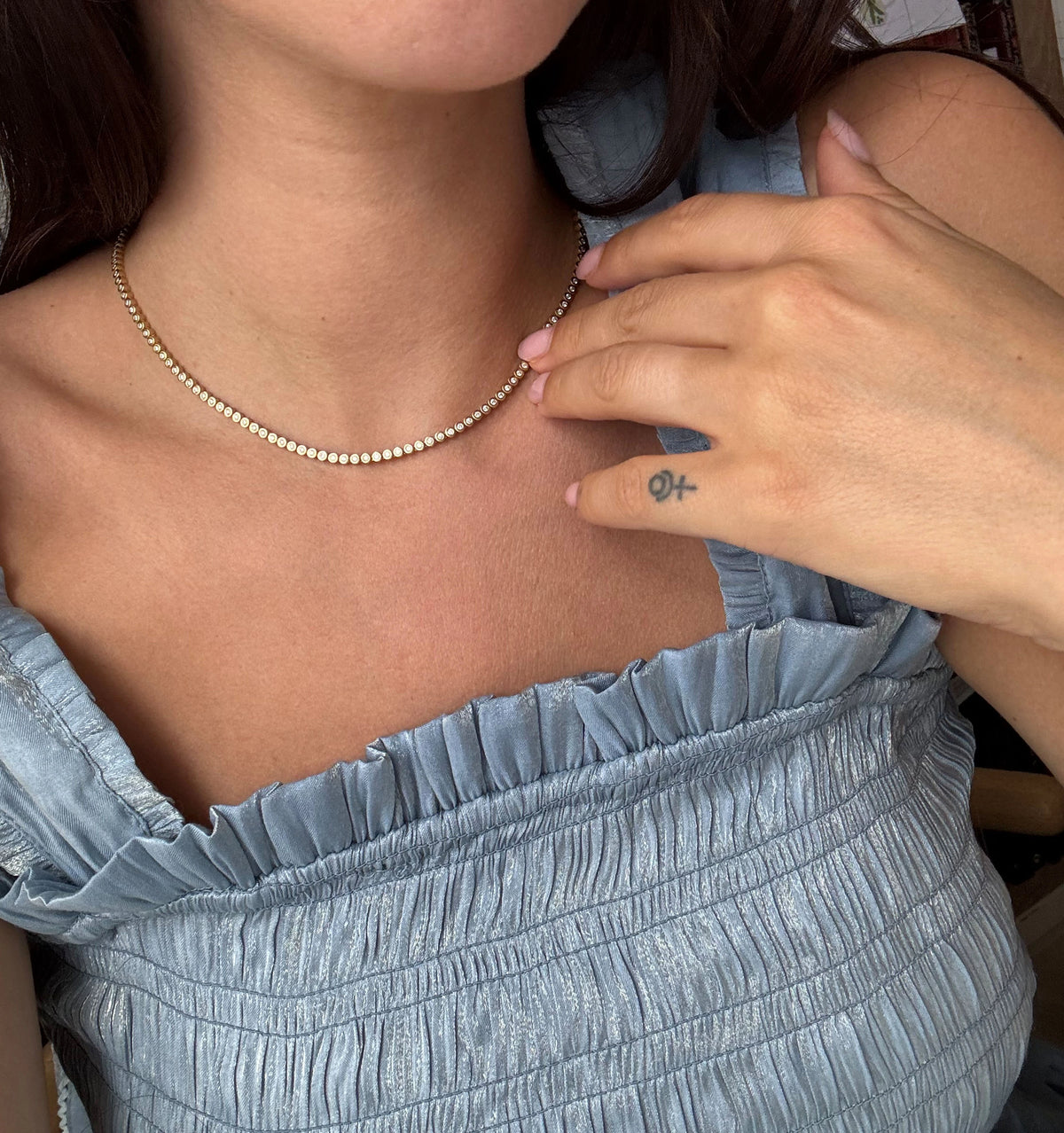 Woman wearing a gold necklace, hand gently touching her collarbone, with a ruffled blue top and a visible tattoo on her hand.