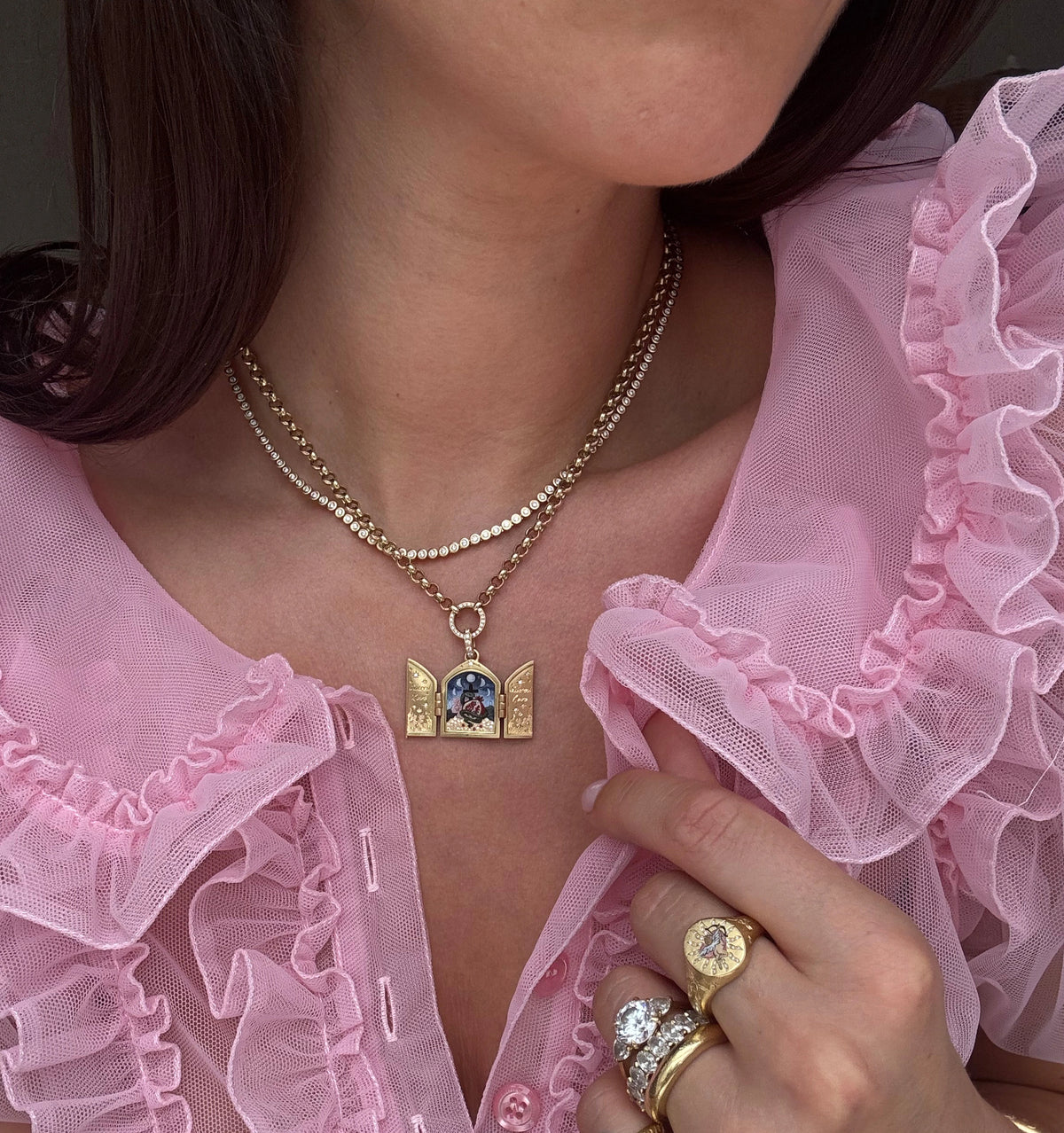 Woman wearing a gold layered necklace with an ornate pendant and multiple golden rings on her fingers.