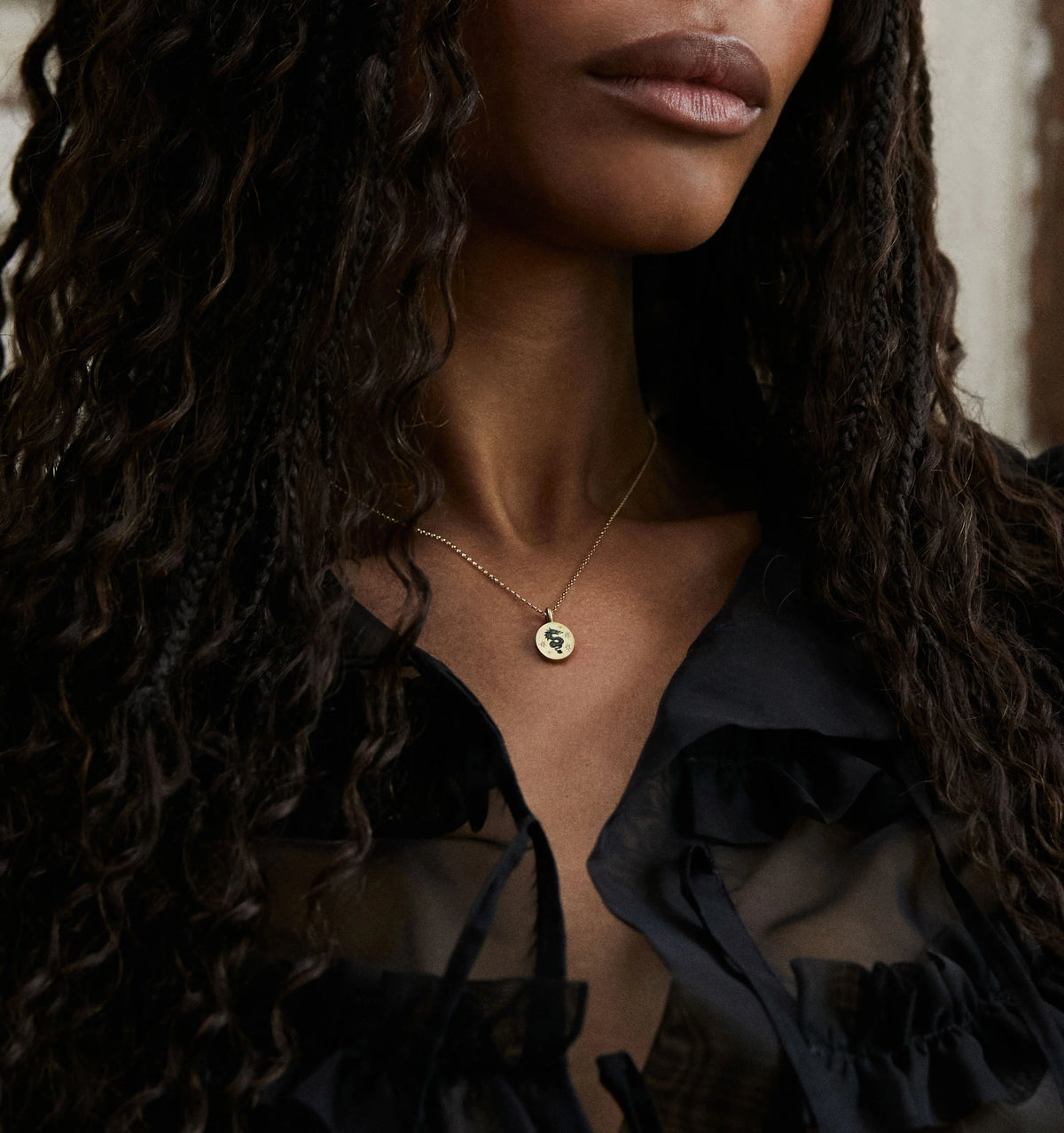 Woman wearing a gold necklace with a circular pendant, styled with a black ruffled blouse.