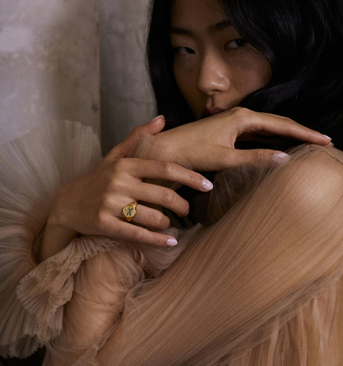 Woman wearing a gold ring with an engraved floral design, hands gently resting on her arm, against a textured backdrop.