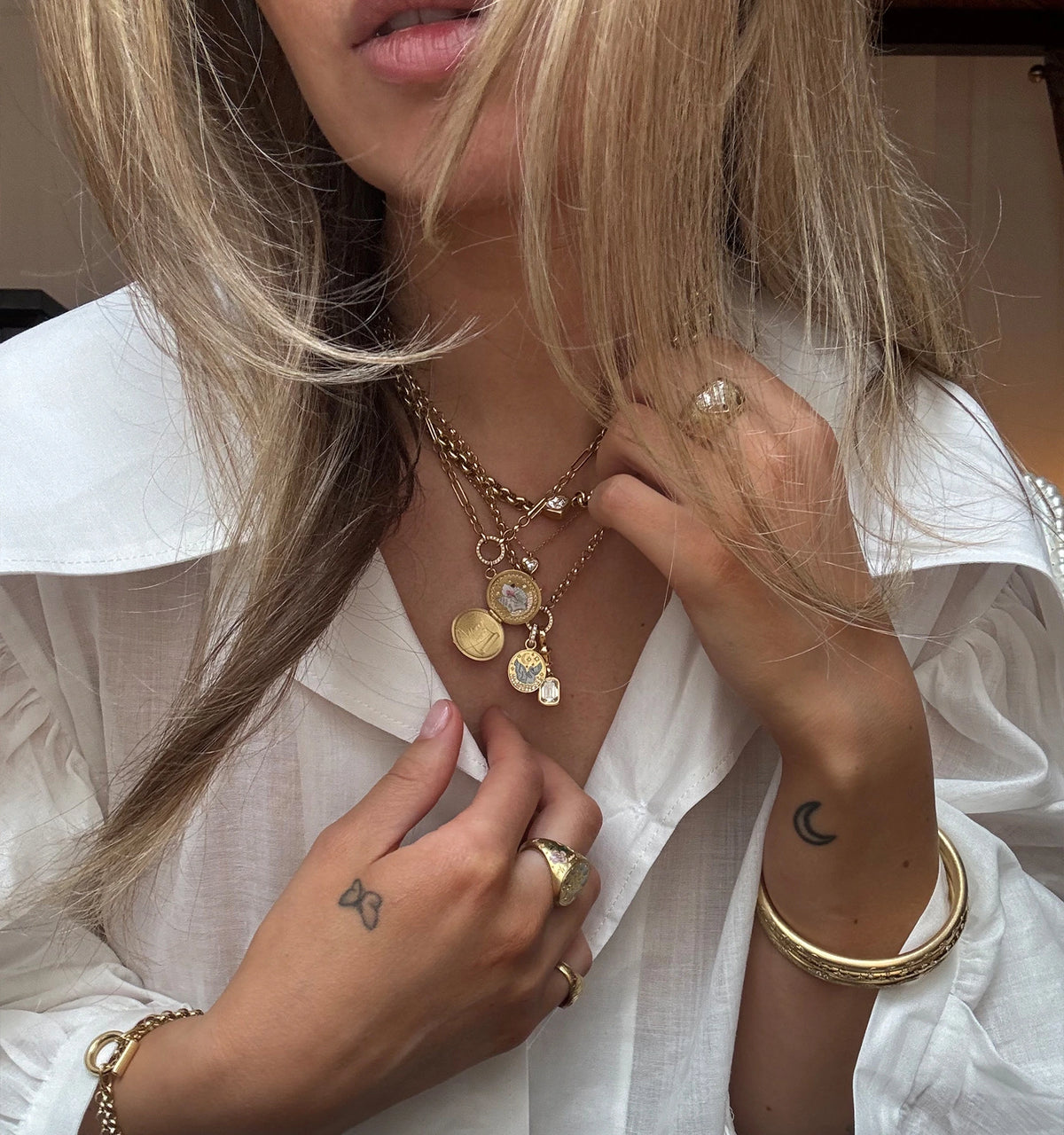 Woman wearing layered gold necklaces with pendants and a gold ring, showing part of a white shirt and tattoos on her hand.