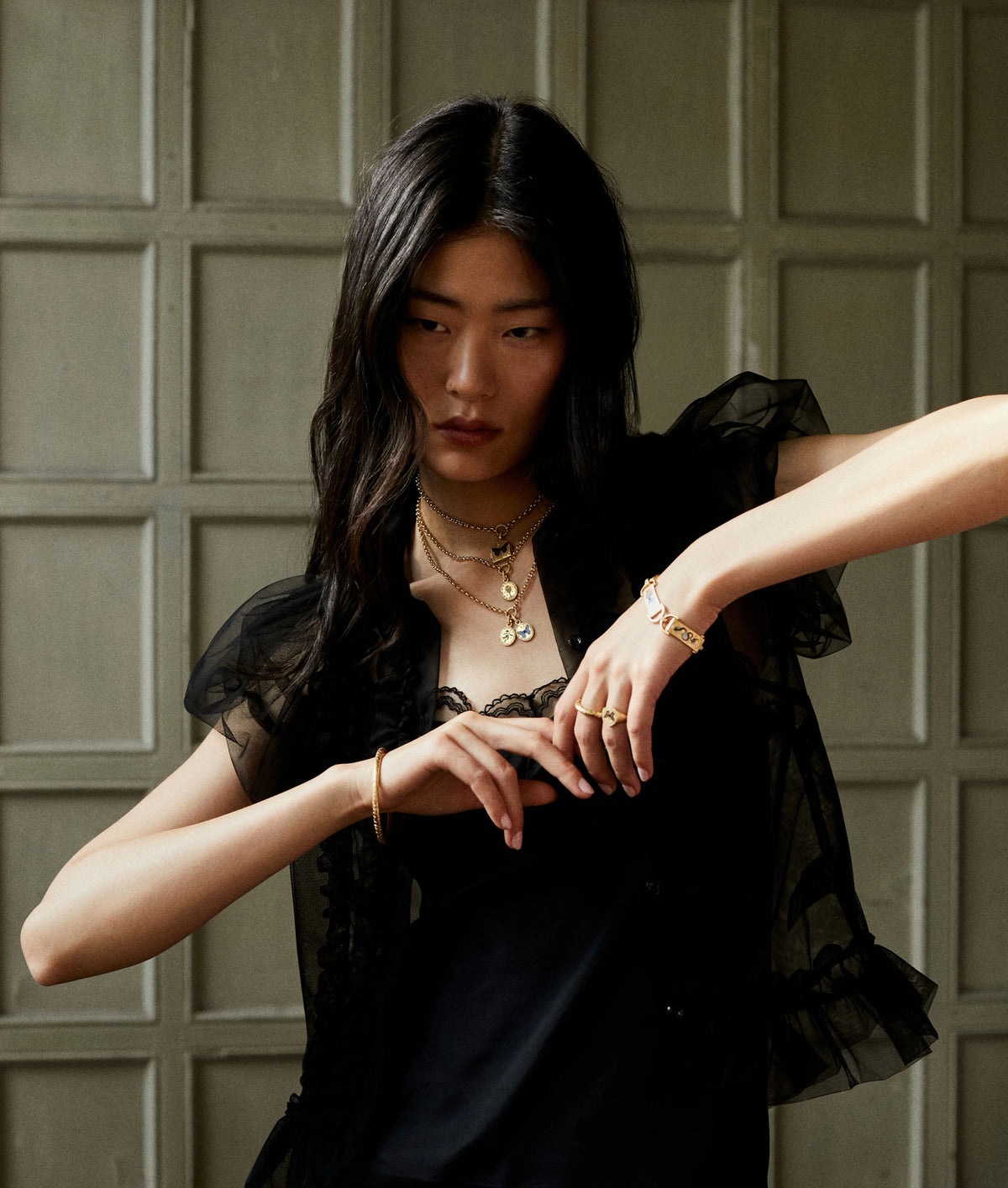 Woman wearing layered gold necklaces and gold rings, paired with a black outfit and sheer overlay.