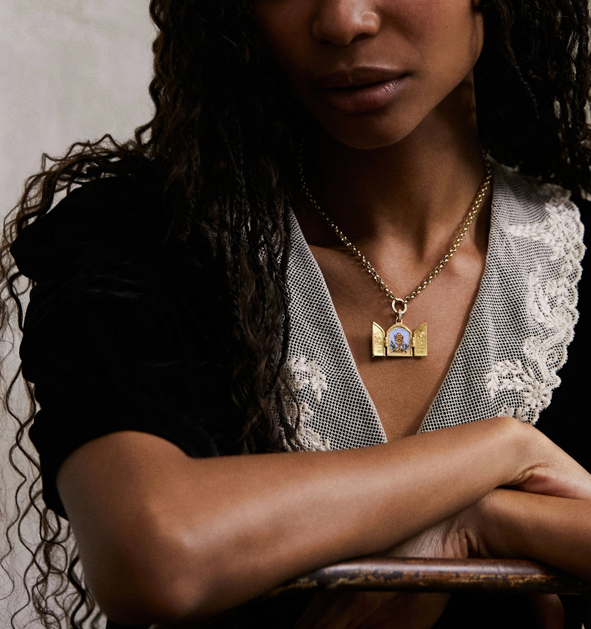 Woman wearing a gold necklace featuring a detailed pendant with an enamel design, resting her arms on a chair.