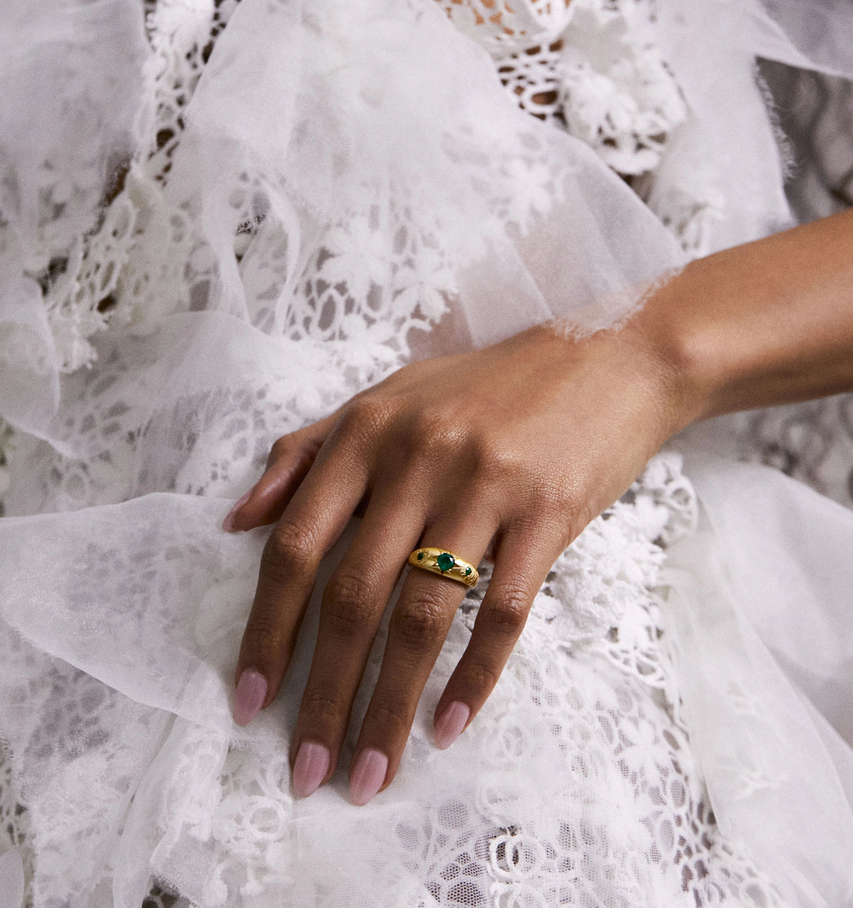 Woman wearing a gold ring set with an emerald, resting on delicate lace fabric.