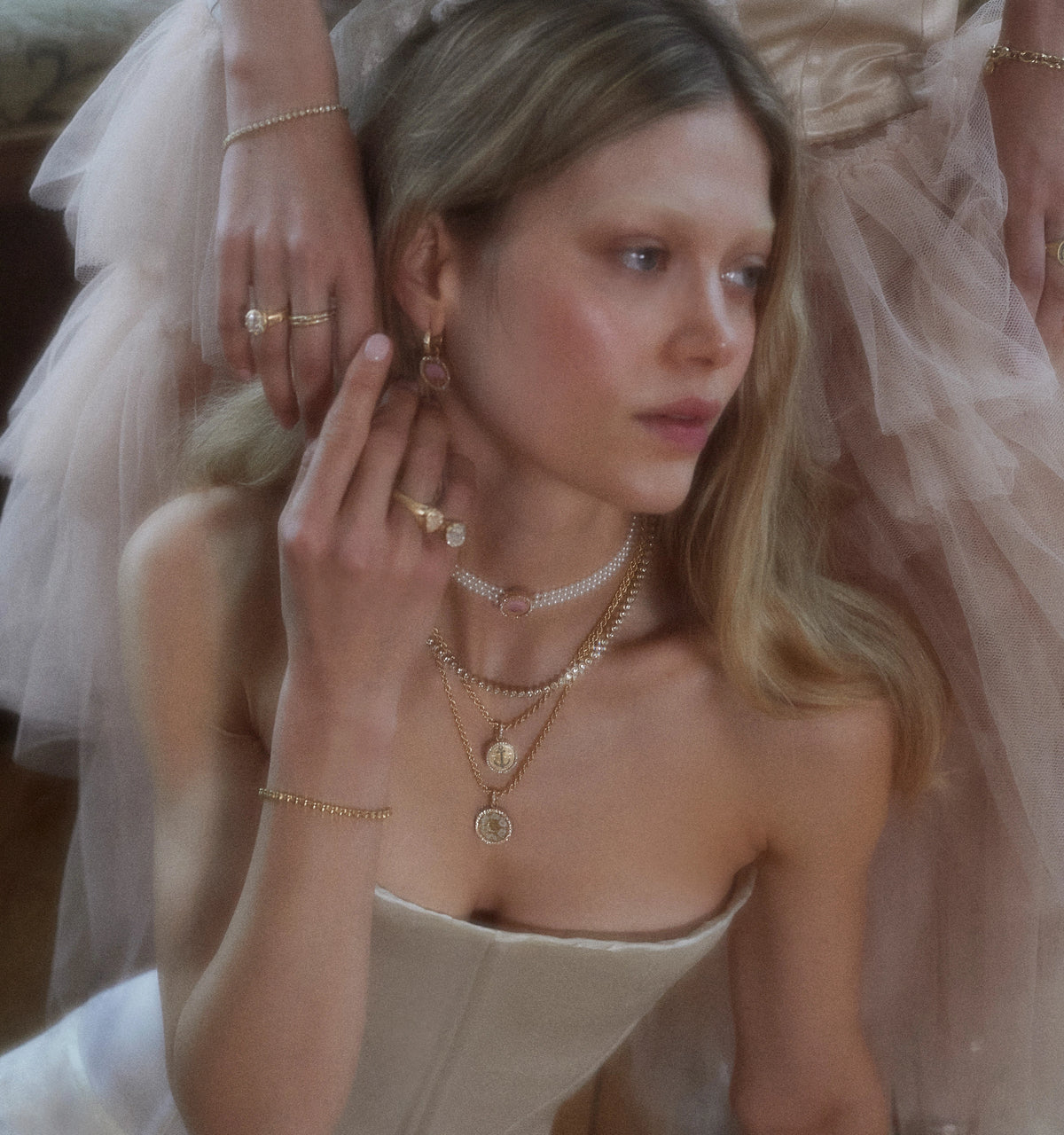 Woman wearing layered gold necklaces with medallion pendants, drop earrings, and rings.
