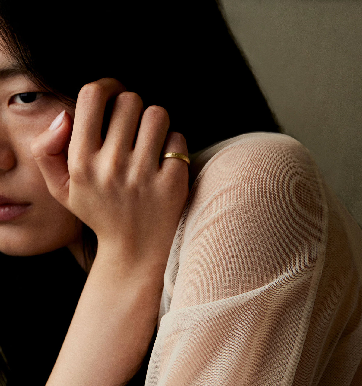 Woman wearing a gold ring, resting her hand on her face, with a sheer light fabric top in a soft-focus background.