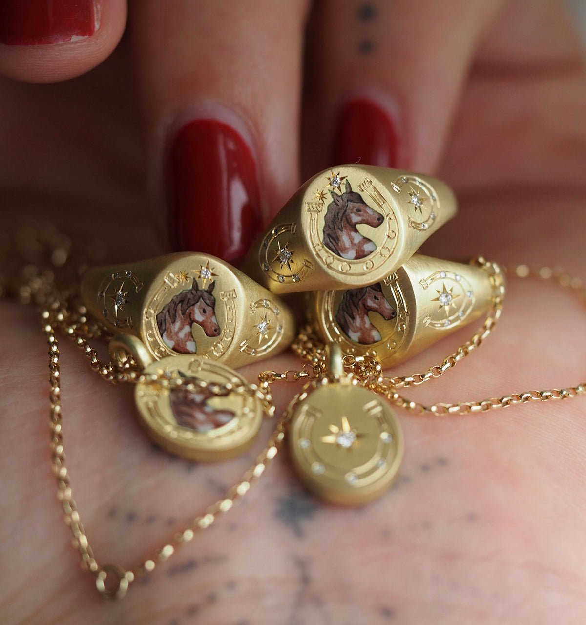 Gold rings and pendants featuring horse motifs on a hand with red nails, resting on a wrist with tattoos.