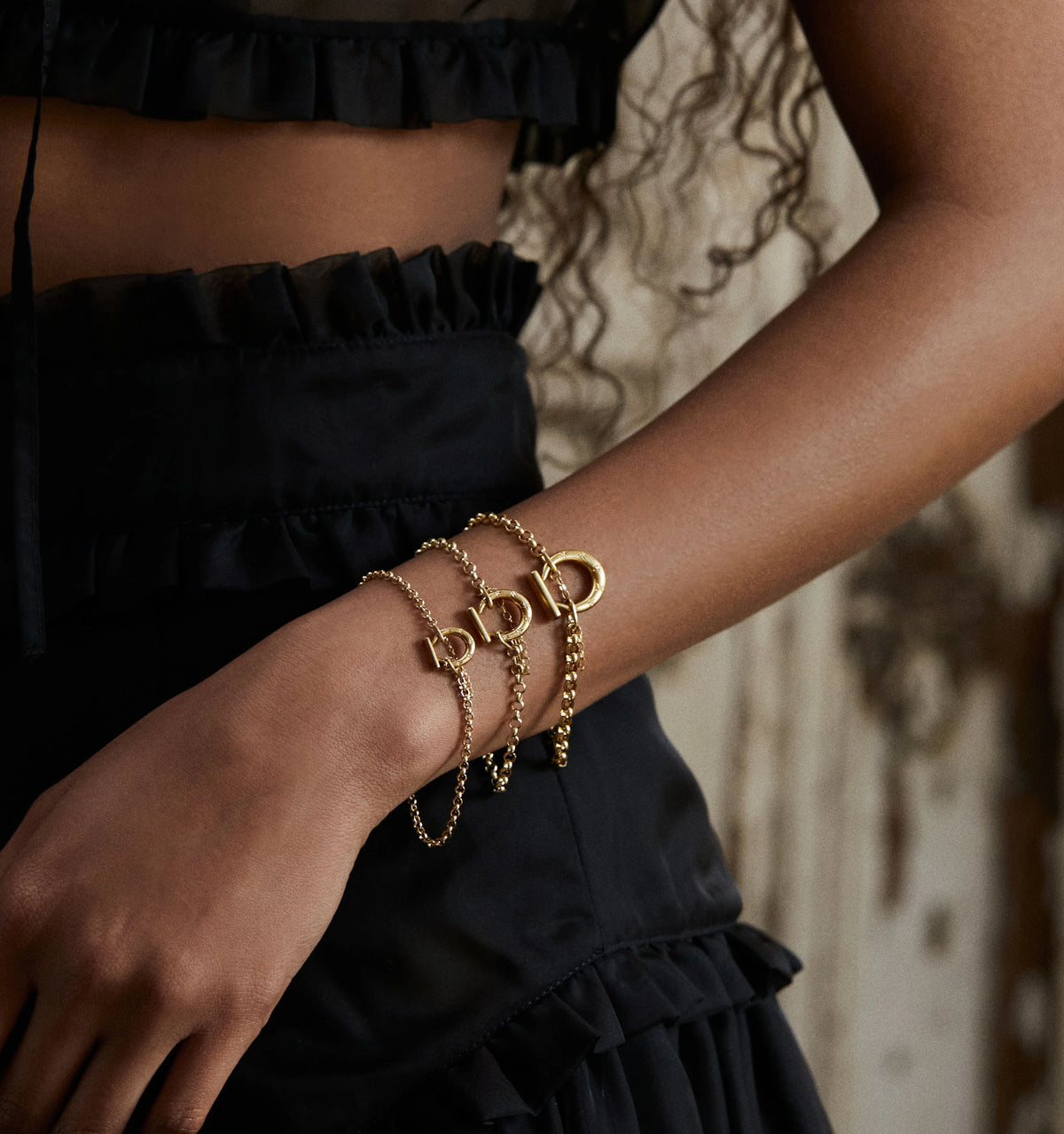 Woman wearing multiple gold bracelets with a toggle clasp, against a textured backdrop.