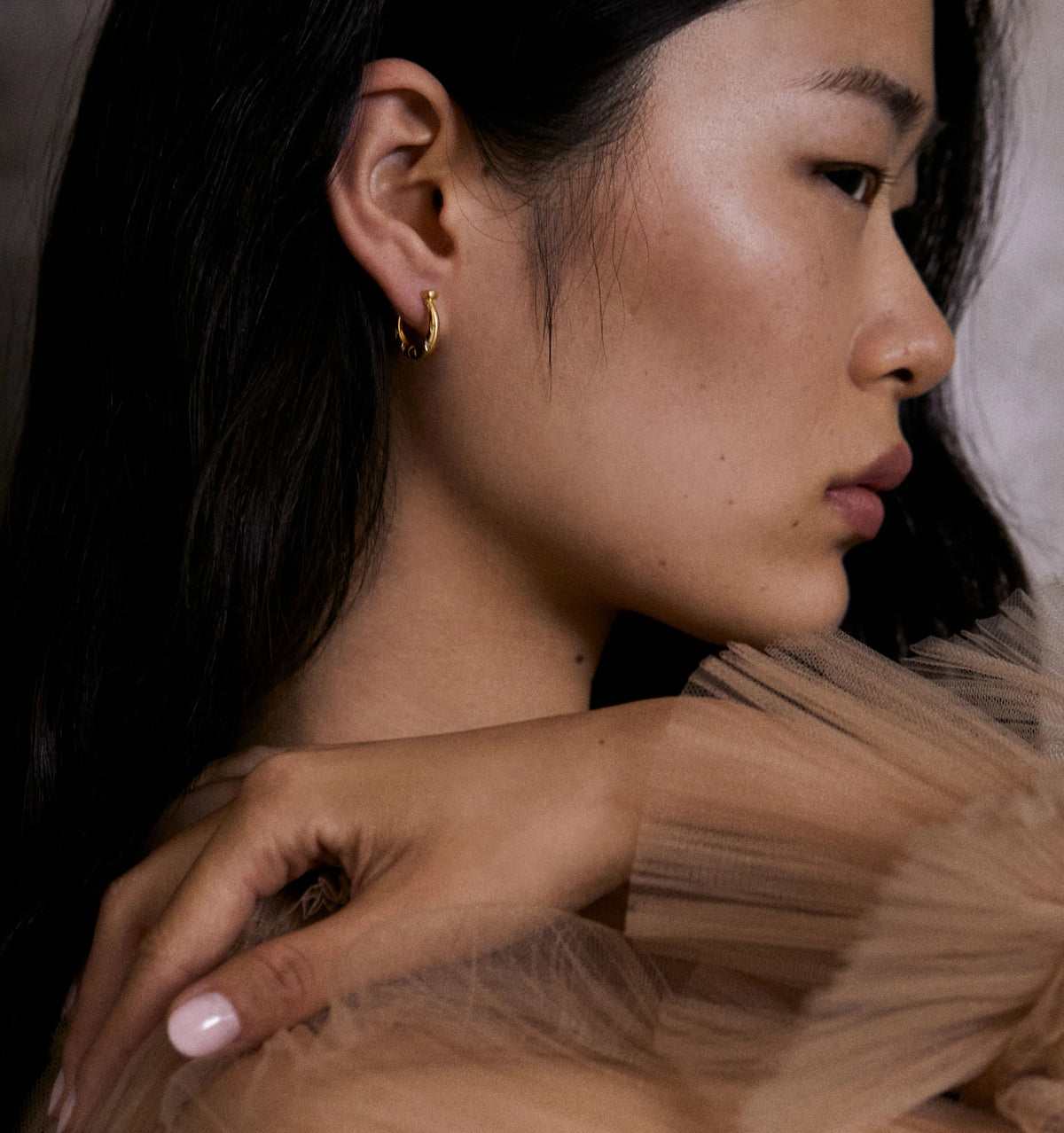 Woman wearing gold hoop earrings, with soft tulle fabric in the background.