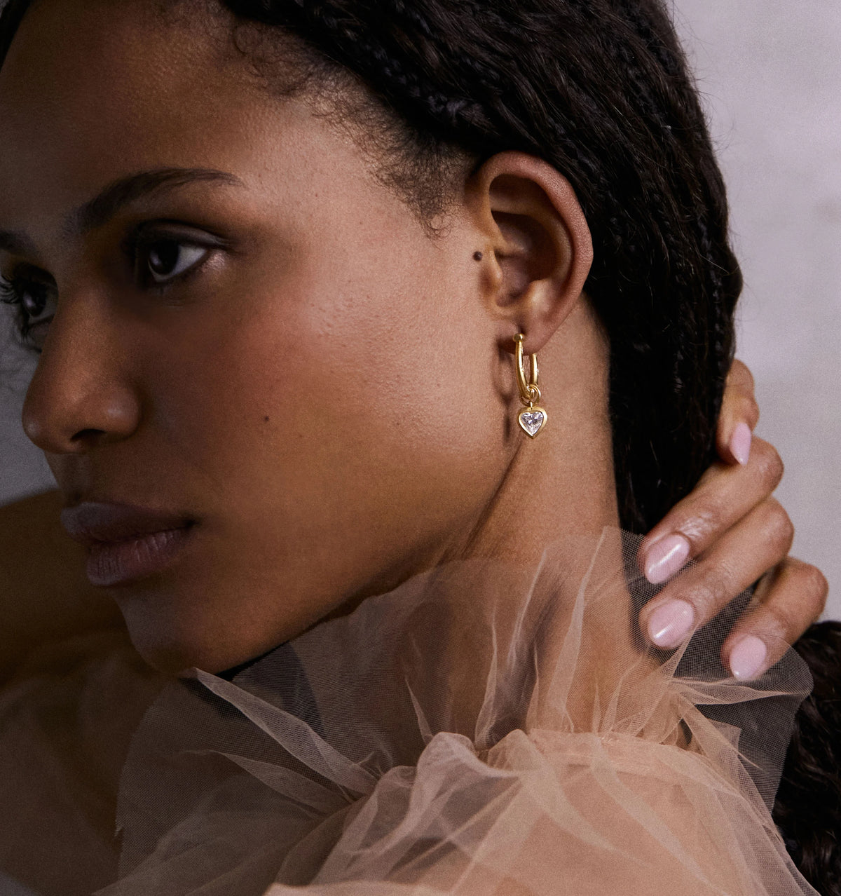 Woman wearing gold hoop earrings with heart-shaped accents, styled with a sheer, ruffled neckline blouse.