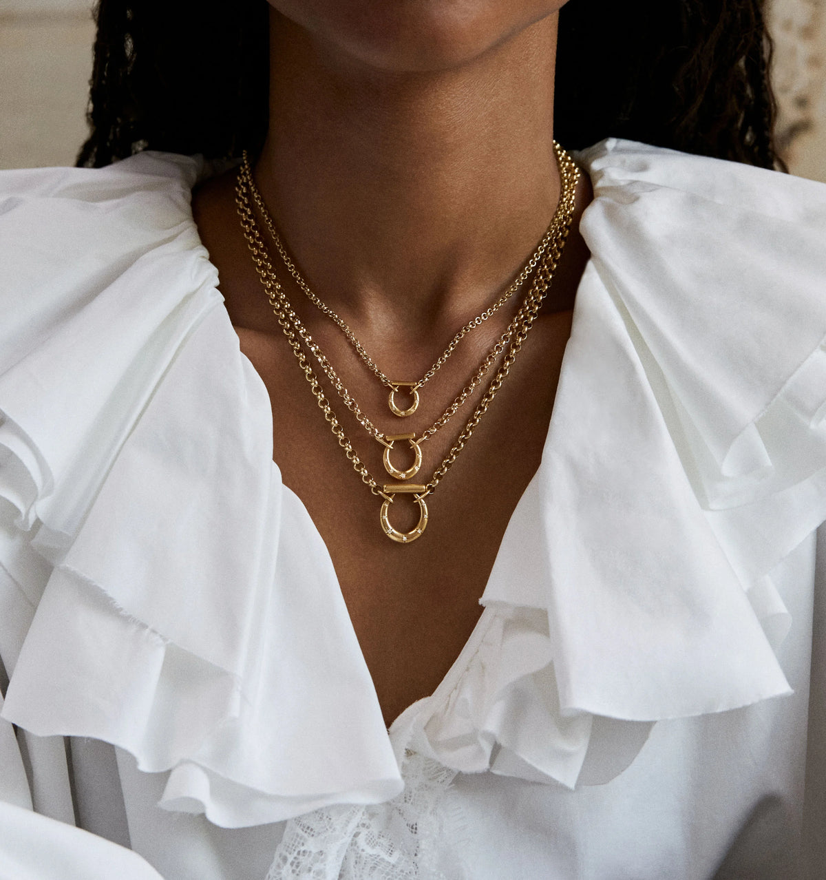 Woman wearing three layered gold necklaces with circular pendants, styled with a white ruffled blouse.